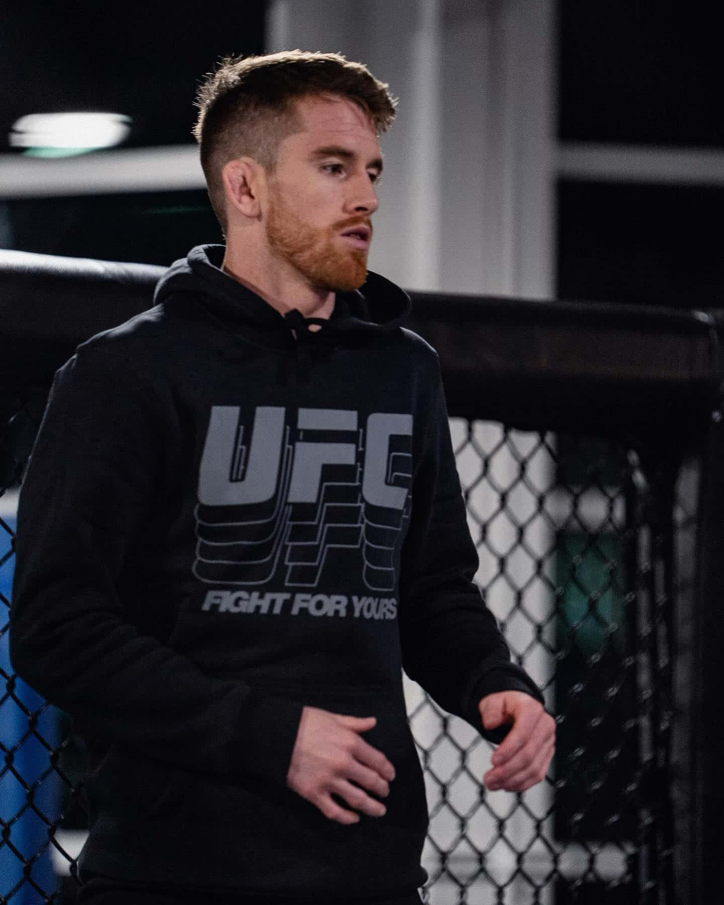 MMA 桌布 壁紙 — 一位專注的男性拳手穿著黑色UFC連帽衫，夜晚站在昏暗的八角籠內。