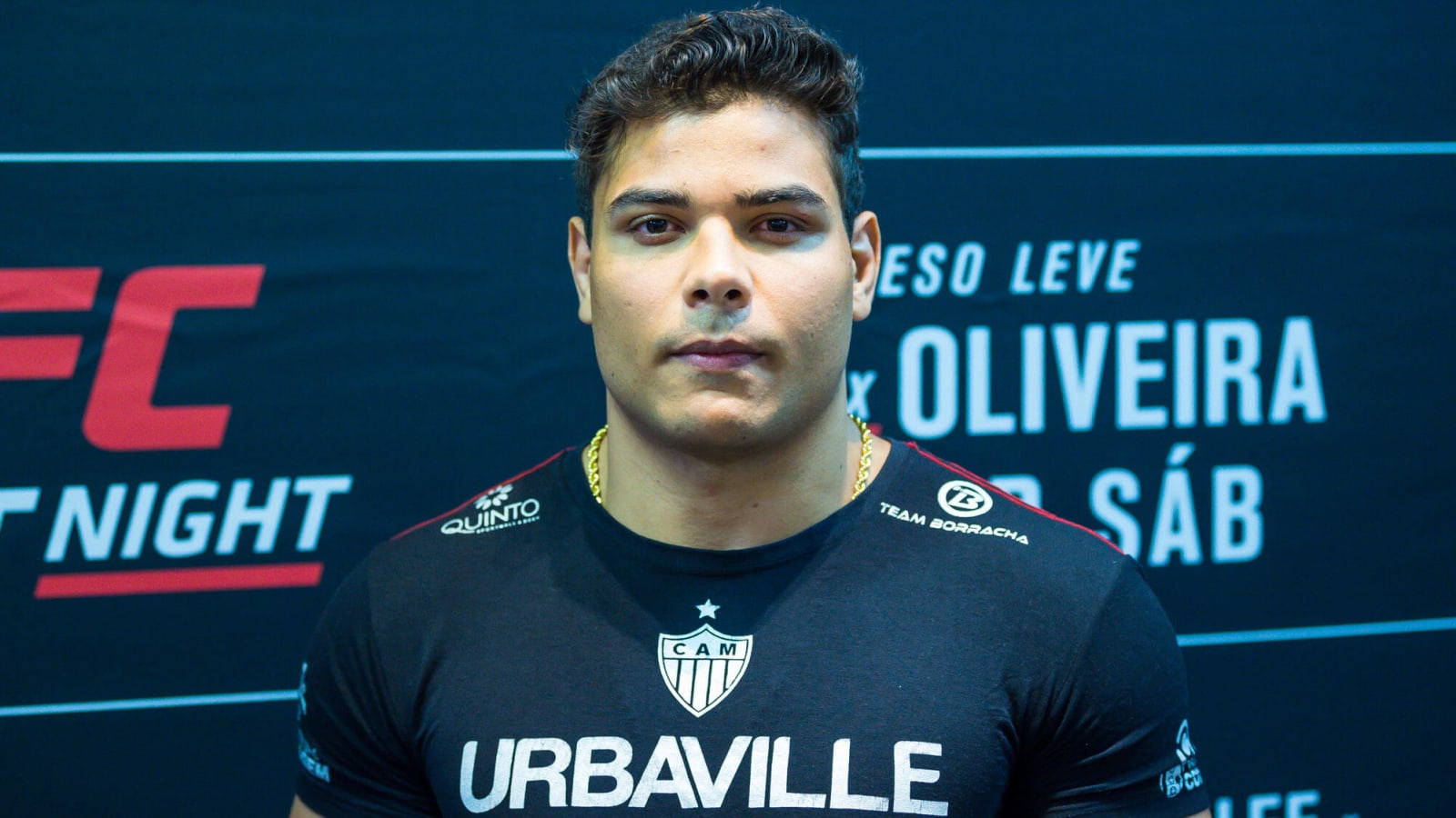 UFC 桌布 壁紙 — 一位專注的男性拳手身穿黑色UFC活動T恤，站在深藍色宣傳背景前，背景上有粗體的紅色和白色文字。