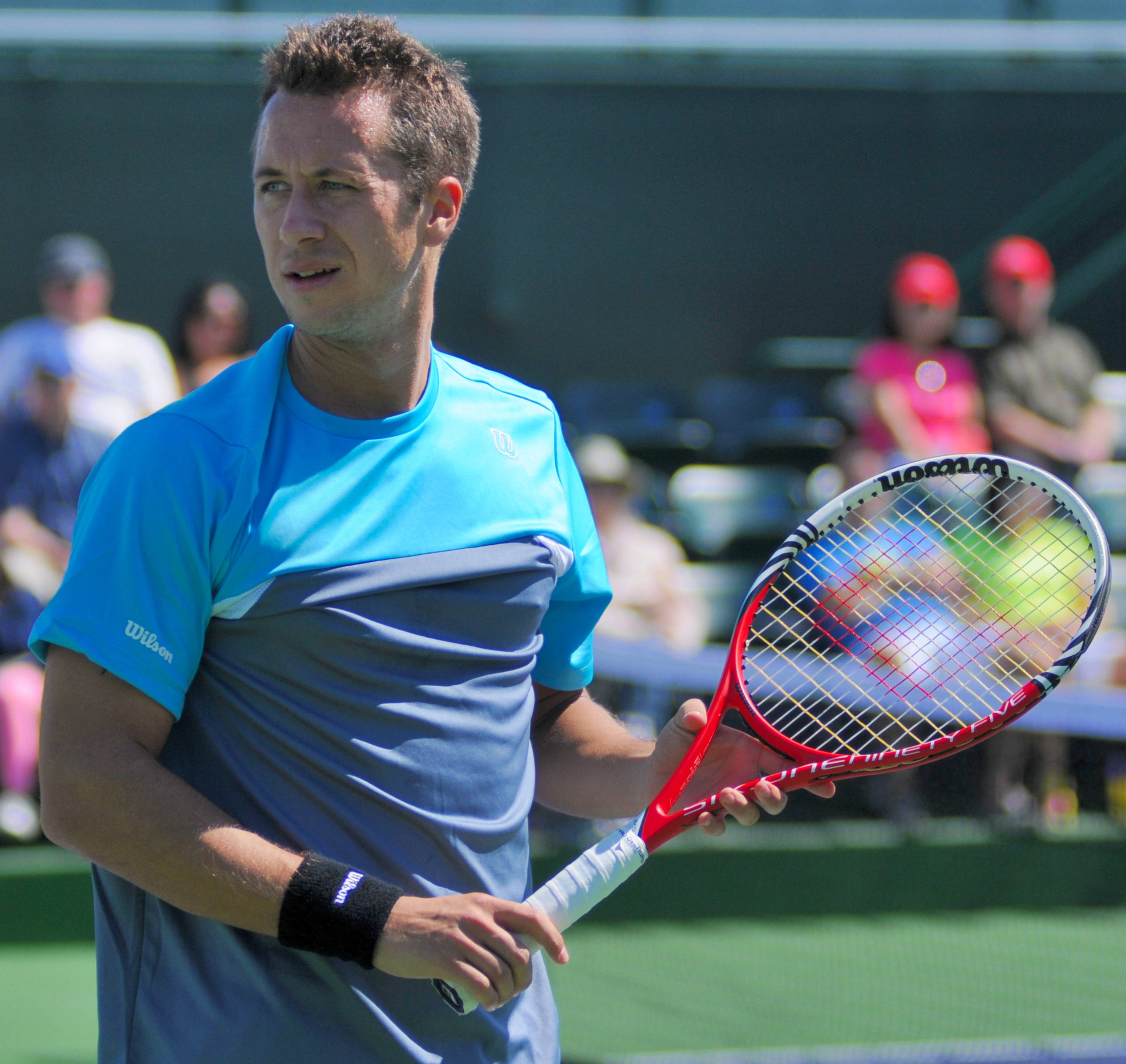 Tennis 桌布 壁紙 — 一位專注的男性網球運動員，身穿藍灰色上衣，在明亮的日光下於綠色球場上握著紅色球拍。