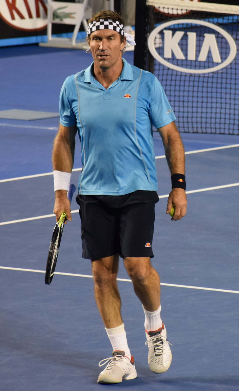 Tennis 桌布 壁紙 — 一位專注的男性網球運動員，身穿藍色襯衫和格子頭帶，在藍色硬地球場上行走，手持球拍和球。