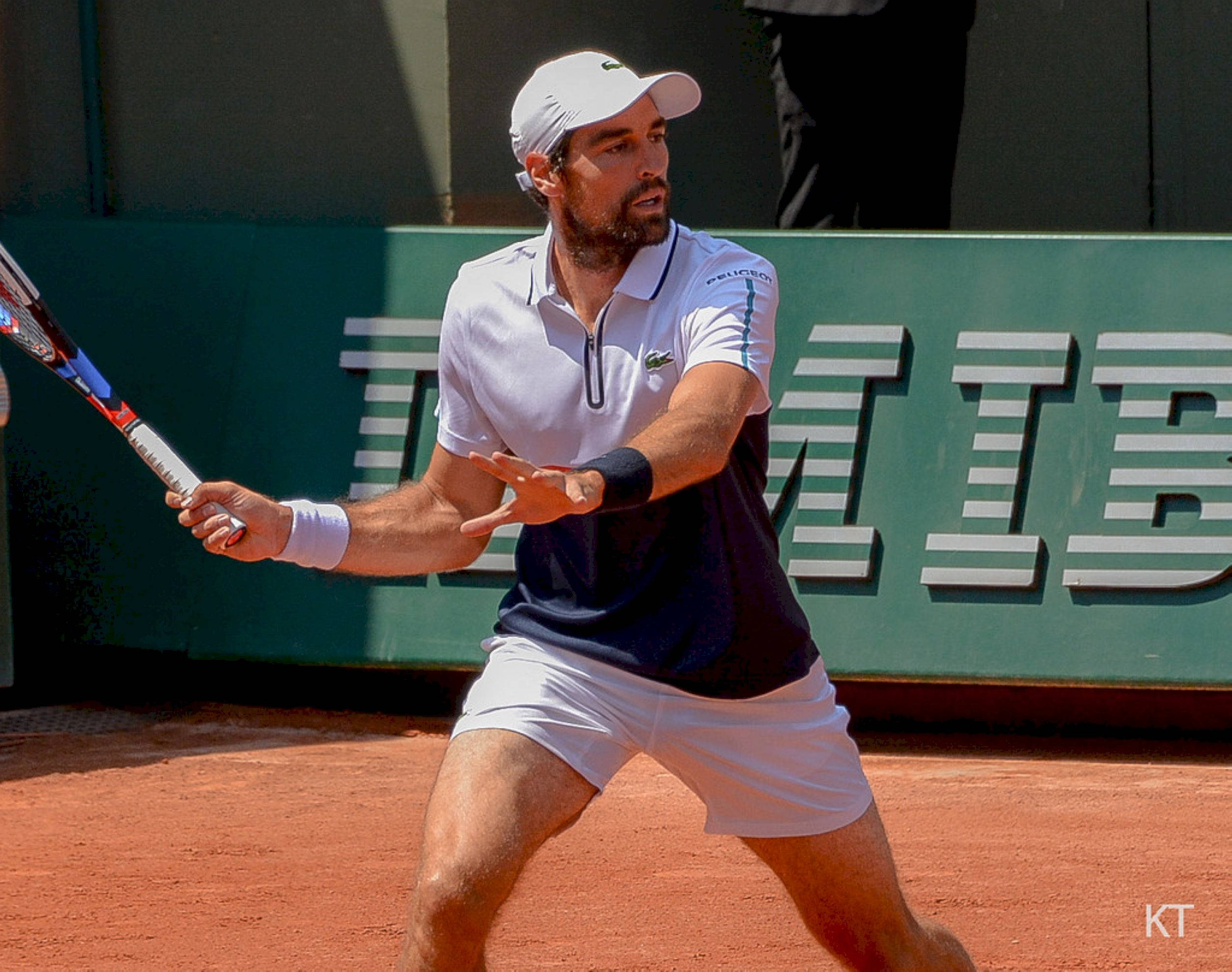 Tennis 桌布 壁紙 — 一位專注的男性網球運動員在陽光普照的紅土球場上揮動球拍，身後有綠色牆壁品牌標誌。