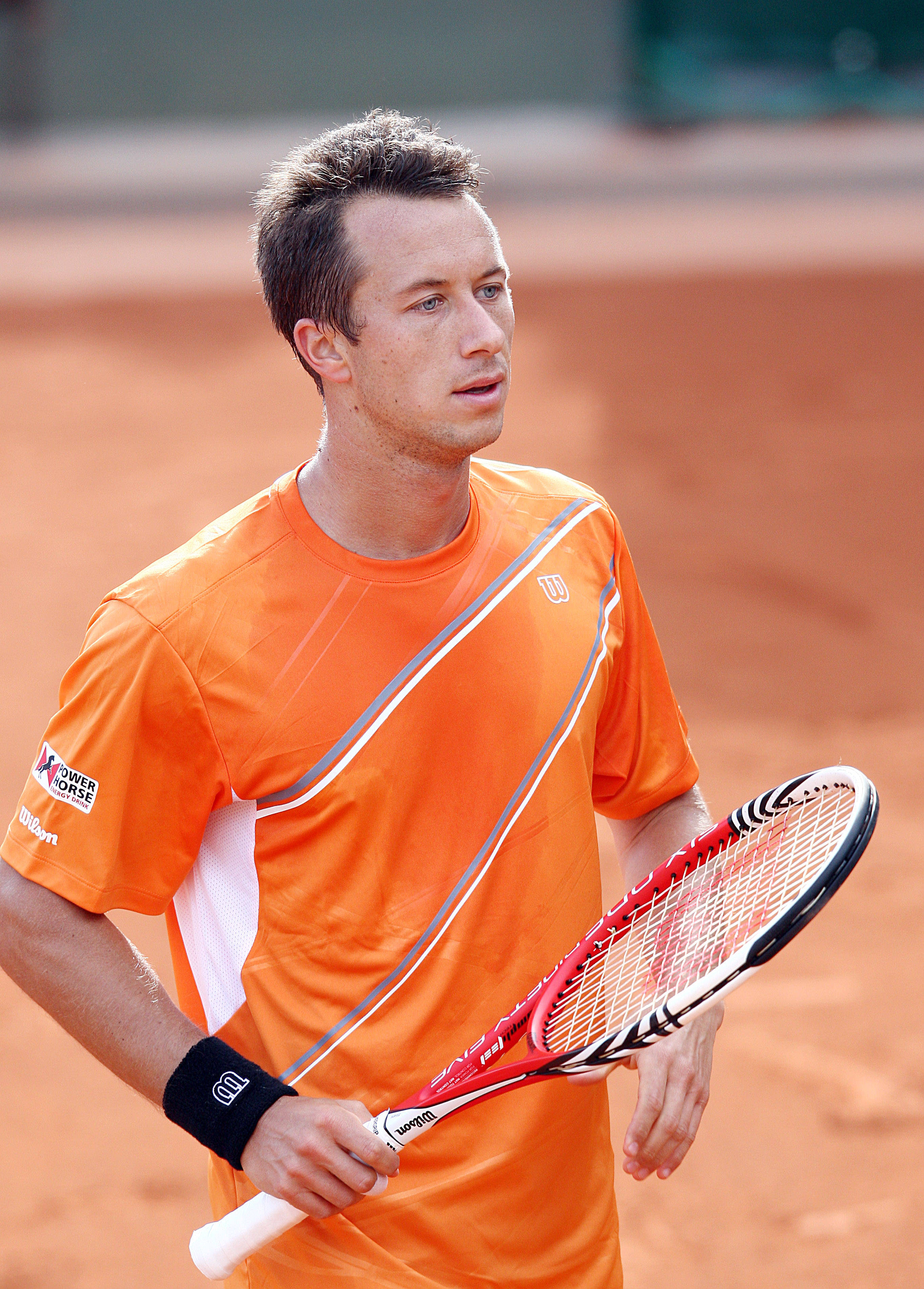 Tennis 桌布 壁紙 — 一位專注的男性網球運動員穿著橙色襯衫，站在紅土球場上，手持紅白相間的球拍。