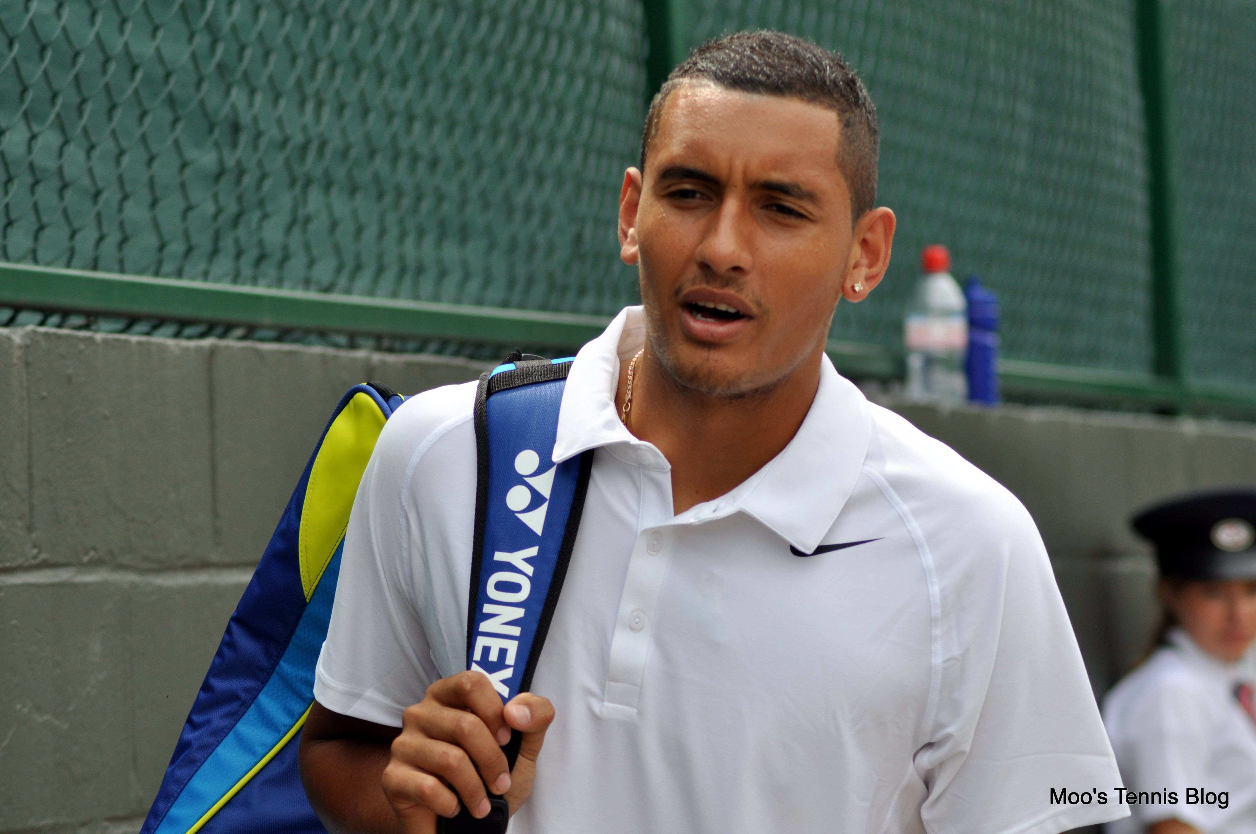 Kyrgios 桌布 壁紙 — 一位專注的男性網球運動員穿著白色Polo衫，在綠色圍欄附近攜帶一個藍黃色的Yonex球拍袋。