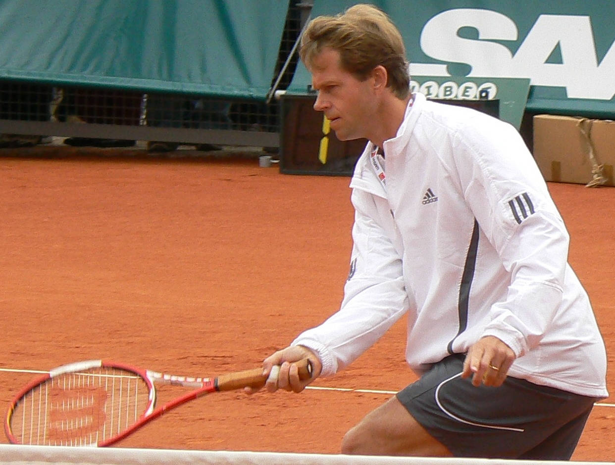 Tennis 桌布 壁紙 — 一位專注的男性網球運動員穿著白色外套，在紅土球場上蹲下，手持紅色球拍。