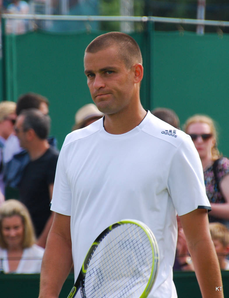 Tennis 桌布 壁紙 — 一位專注的男性網球運動員身穿白色adidas球衣，在草地場上握著球拍，背景有觀眾。
