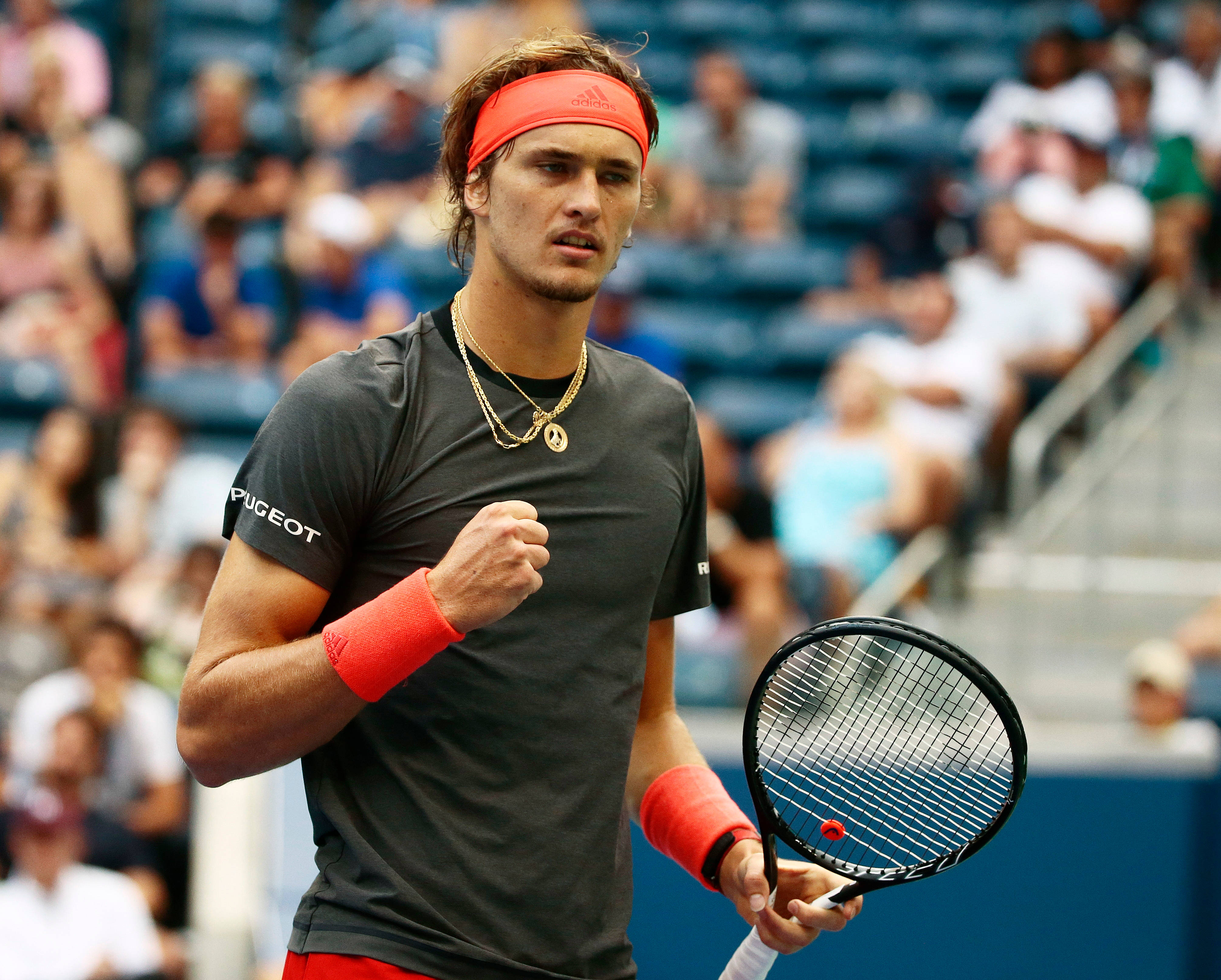 alexander zverev 桌布 壁紙 — 一位專注的男性網球選手，戴著紅色頭帶和護腕，在陽光明媚的球場上比賽中握緊拳頭，背景是模糊的觀眾。