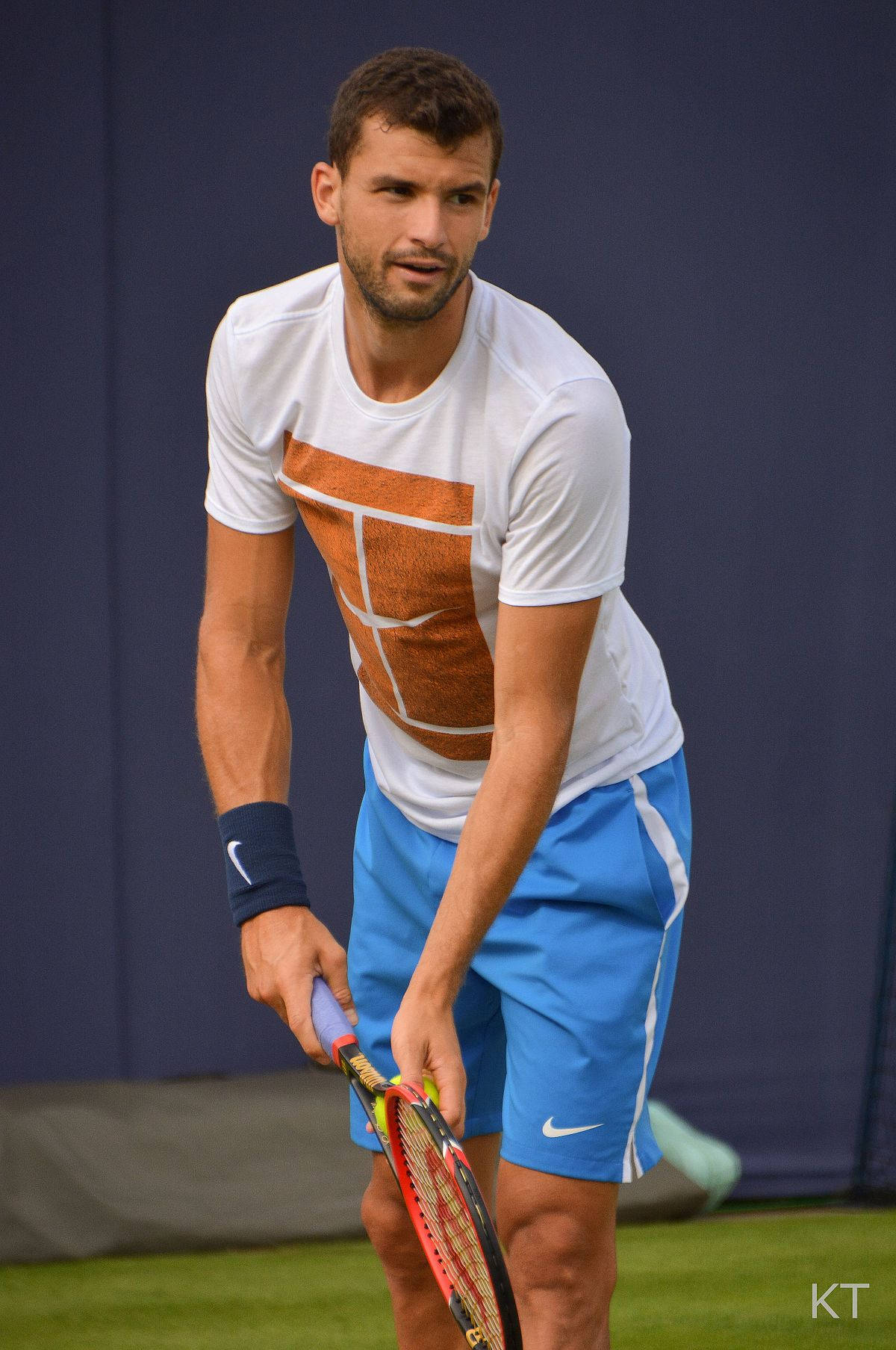 Tennis 桌布 壁紙 — 一位專注的男性網球選手，身穿白色和藍色裝備，在綠色球場上握著球拍，背景是深藍色牆壁。