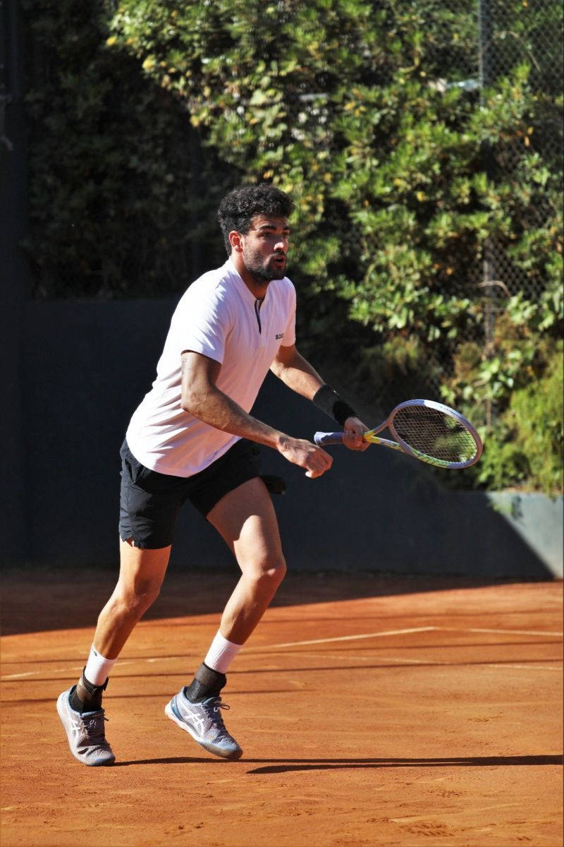 Tennis 桌布 壁紙 — 一位專注的男性網球選手，身穿白色襯衫和黑色短褲，在陽光明媚的紅土球場上向前猛衝，周圍環繞著綠色樹葉。