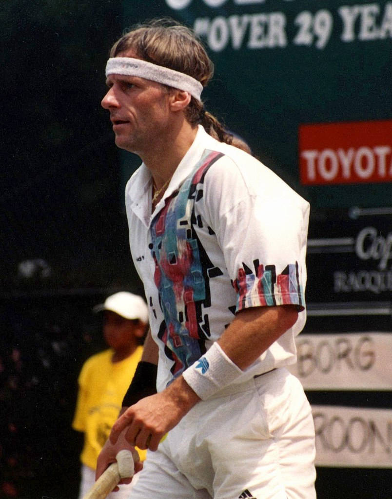 Tennis 桌布 壁紙 — 一位專注的男性網球選手，身穿色彩繽紛的80年代襯衫和頭帶，在帶有贊助商橫幅的球場上比賽中握緊球拍。