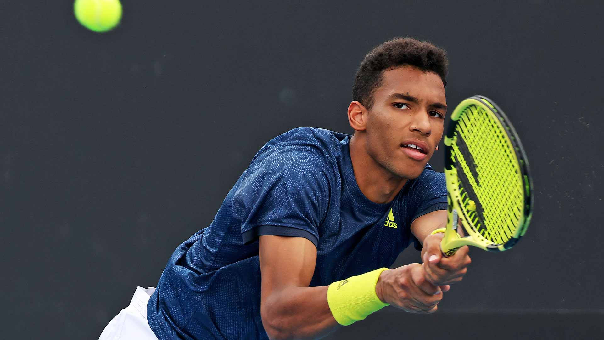 felix auger aliassime 桌布 壁紙 — 一位專注的男性網球選手，身穿藍色襯衫和螢光黃色腕帶，在深色背景下揮拍擊打亮綠色球。