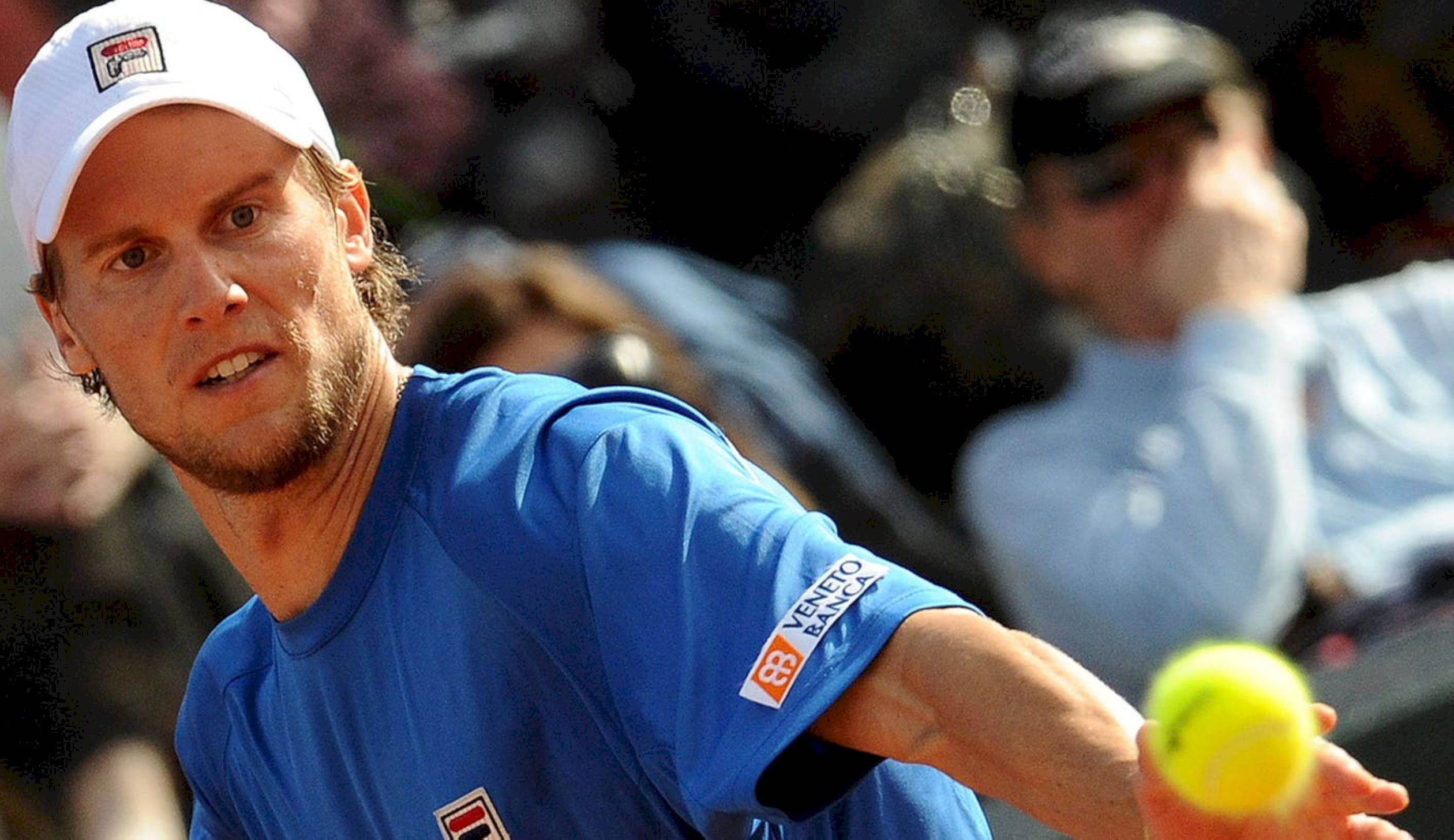 Tennis 桌布 壁紙 — 一位專注的男性網球選手，身穿藍色襯衫和白色帽子，準備在紅土球場上發出一顆亮黃色的網球。