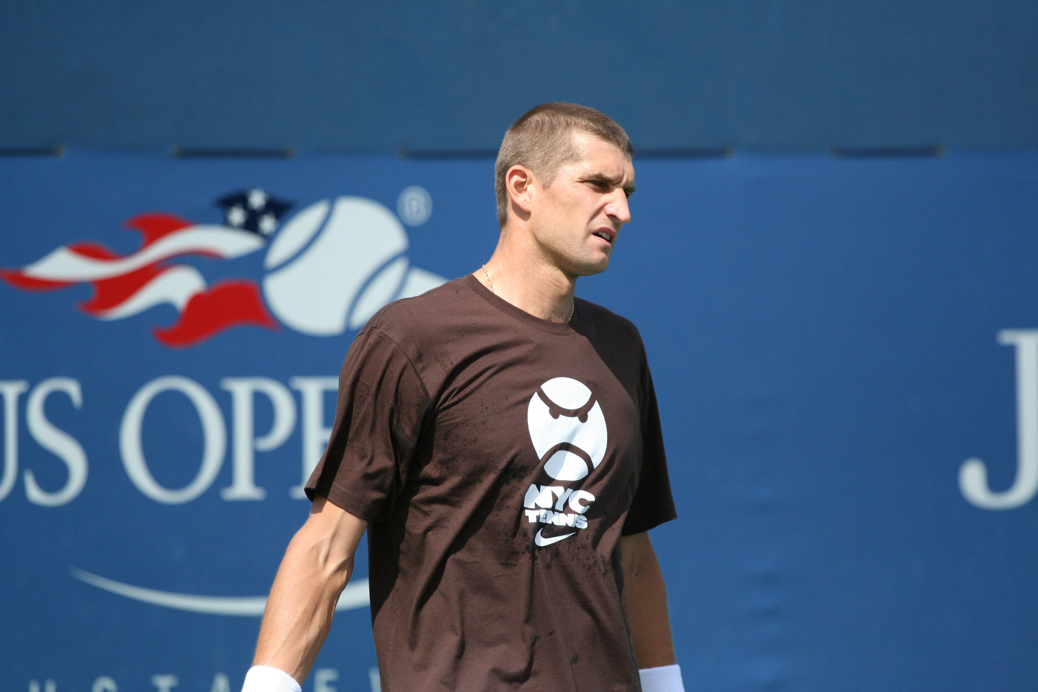 運動 桌布 壁紙 — 一位專注的男性網球選手身穿棕色NYC Tennis襯衫，站在藍色US Open背景的球場上。