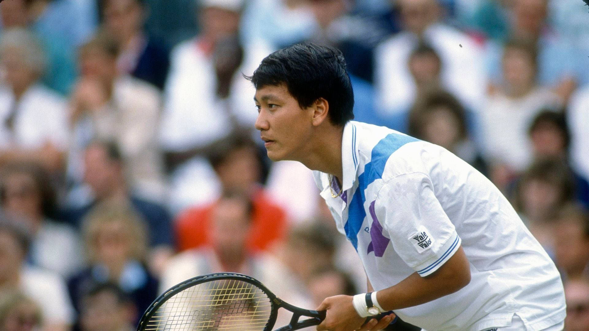 Tennis 桌布 壁紙 — 一位專注的男性網球選手身穿白色和藍色裝備，在比賽中緊握球拍，背景有模糊的觀眾。