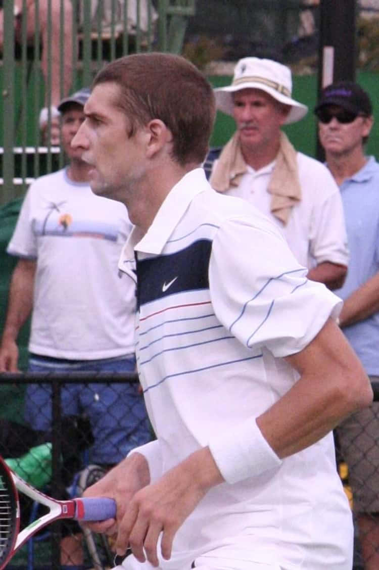 Athlete 桌布 壁紙 — 一位專注的男性網球選手身穿白色裝備，在比賽中緊握球拍，觀眾在圍欄後觀看。