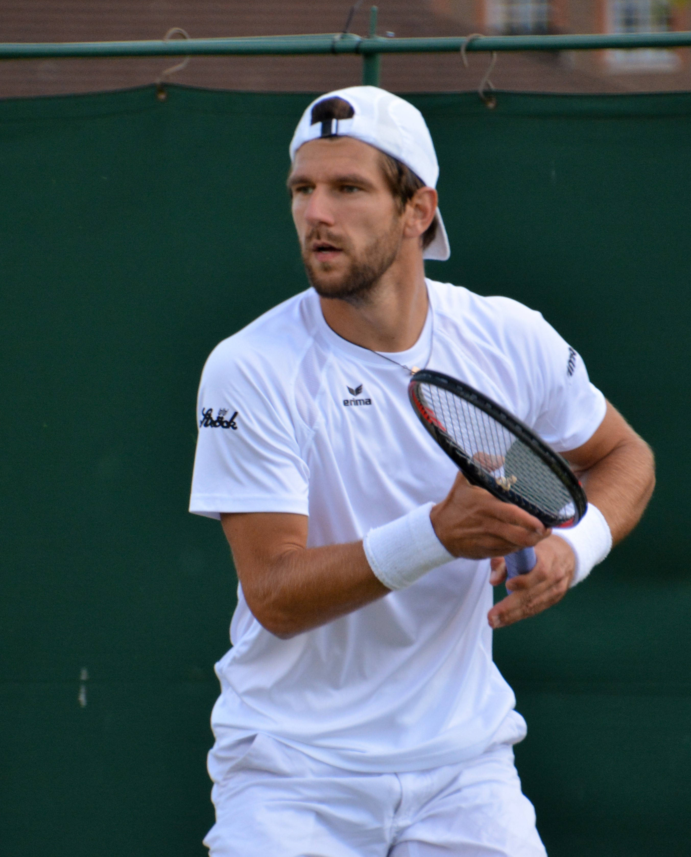 Tennis 桌布 壁紙 — 一位專注的男性網球選手身穿白色裝備，在比賽中握著球拍，背景是綠色球場圍欄。