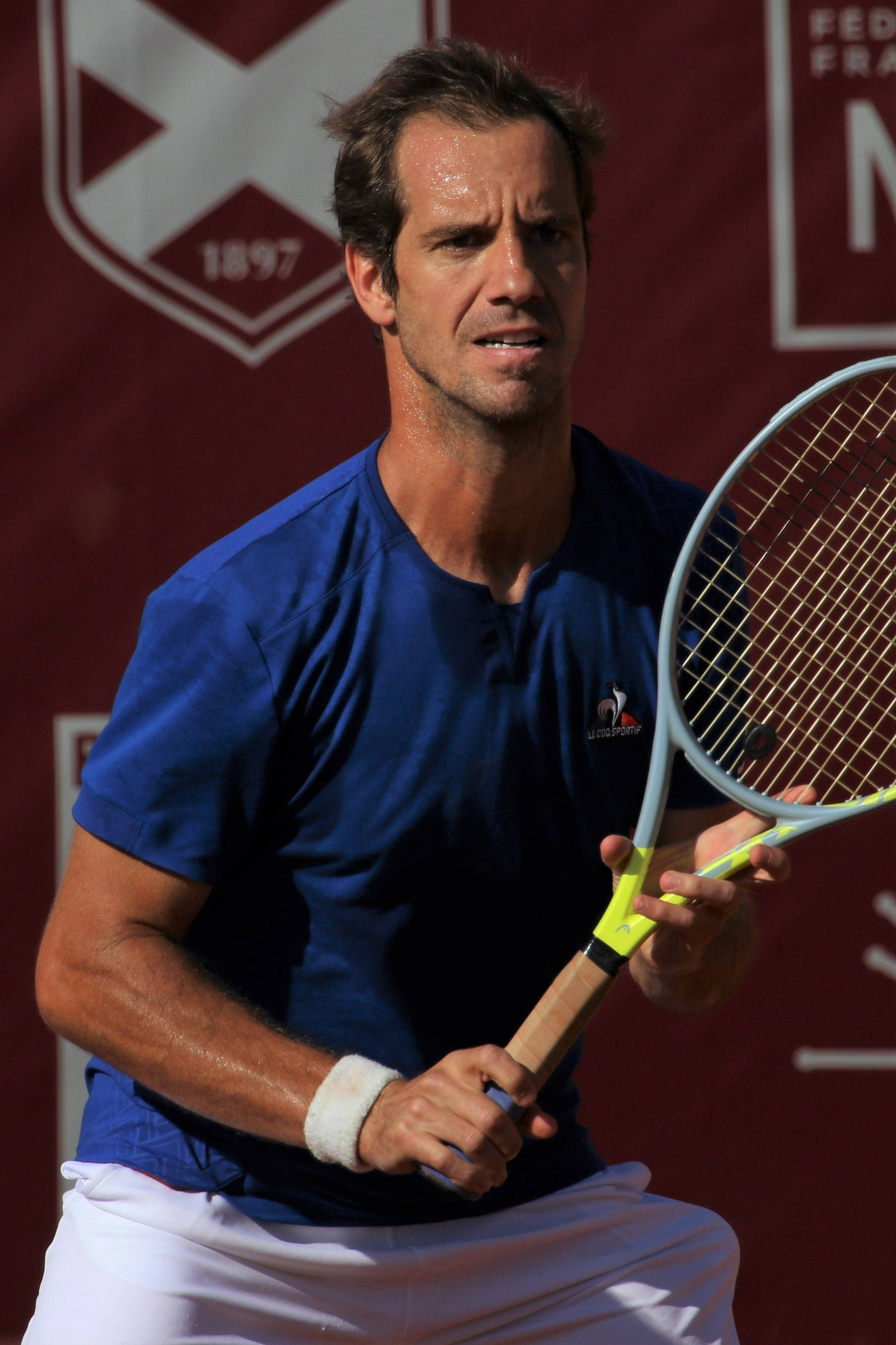 Tennis 桌布 壁紙 — 一位專注的男性網球選手身穿藍色襯衫和白色短褲，在深紅色背景下比賽中緊握球拍。