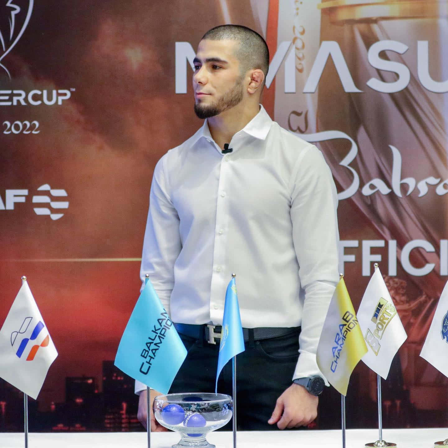 MMA 桌布 壁紙 — 一位專注的男性運動員穿著白襯衫，站在一張放有錦標賽旗幟和獎盃碗的桌子旁，參加正式活動。