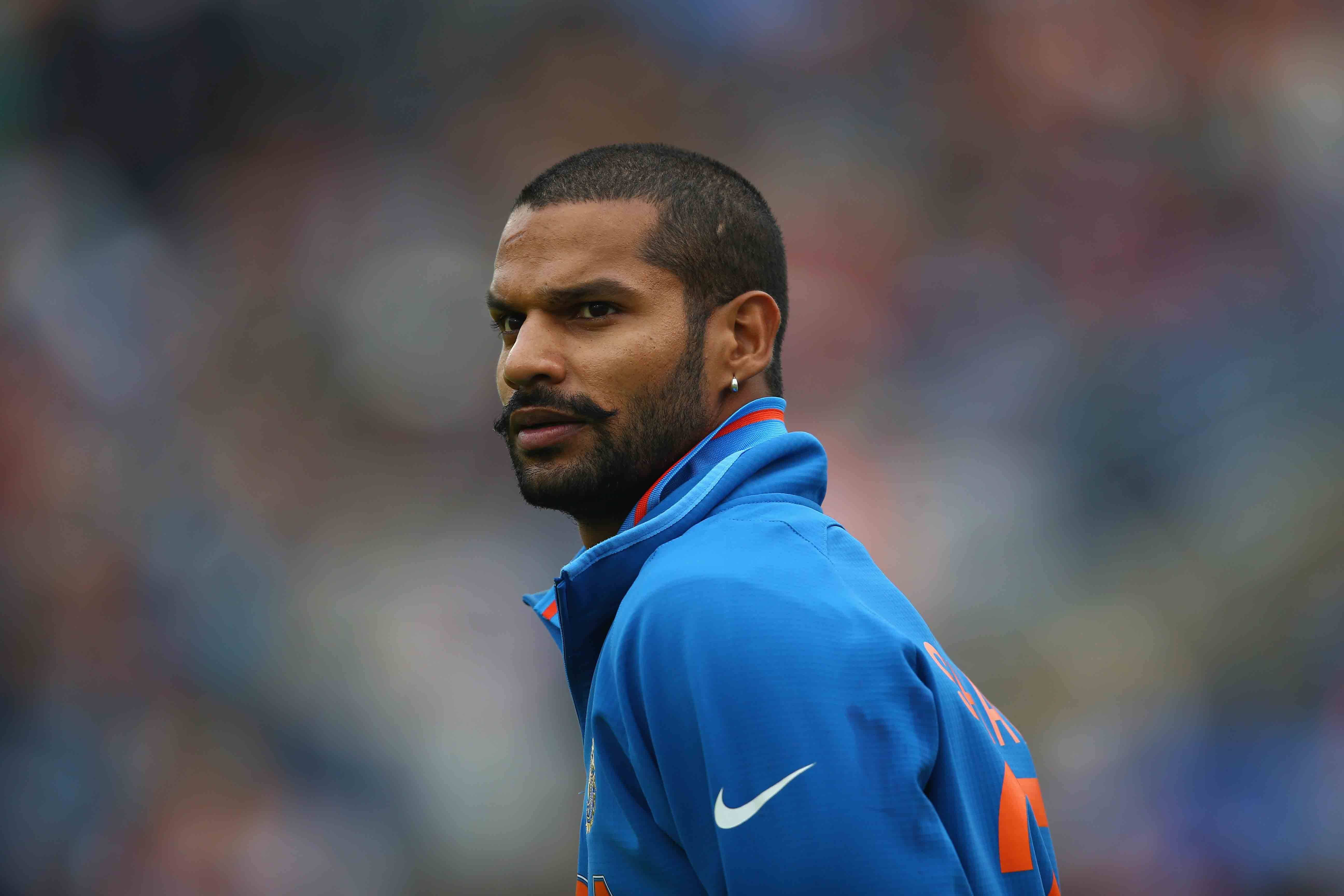shikhar dhawan 桌布 壁紙 — 一位專注的男性運動員身穿藍色運動外套，在模糊的體育場人群中凝視著。