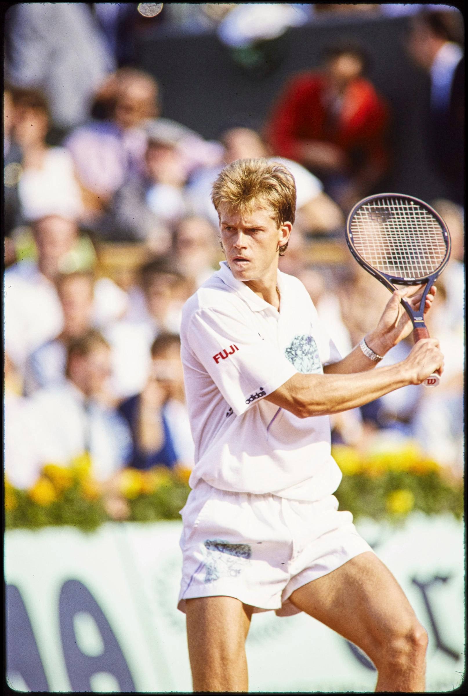 Tennis 桌布 壁紙 — 一位專注的男網球運動員穿著白色裝備，在陽光明媚的戶外球場上比賽中揮拍，背景有模糊的觀眾。