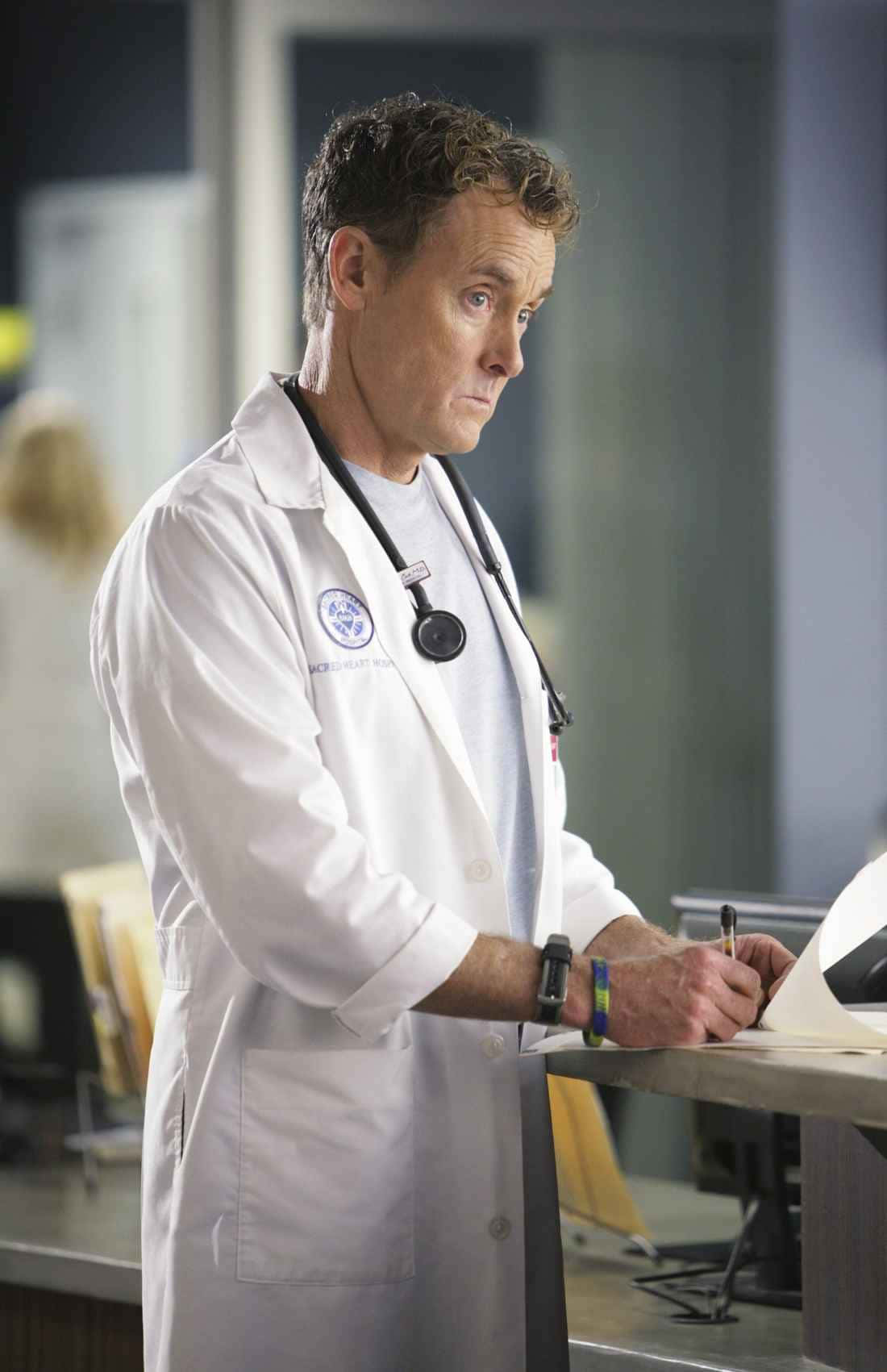 John C. Mcginley 桌布 壁紙 — 一位專注的男醫生穿著白色實驗室外套和聽診器，在明亮的臨床燈光下於醫院桌上專心書寫。