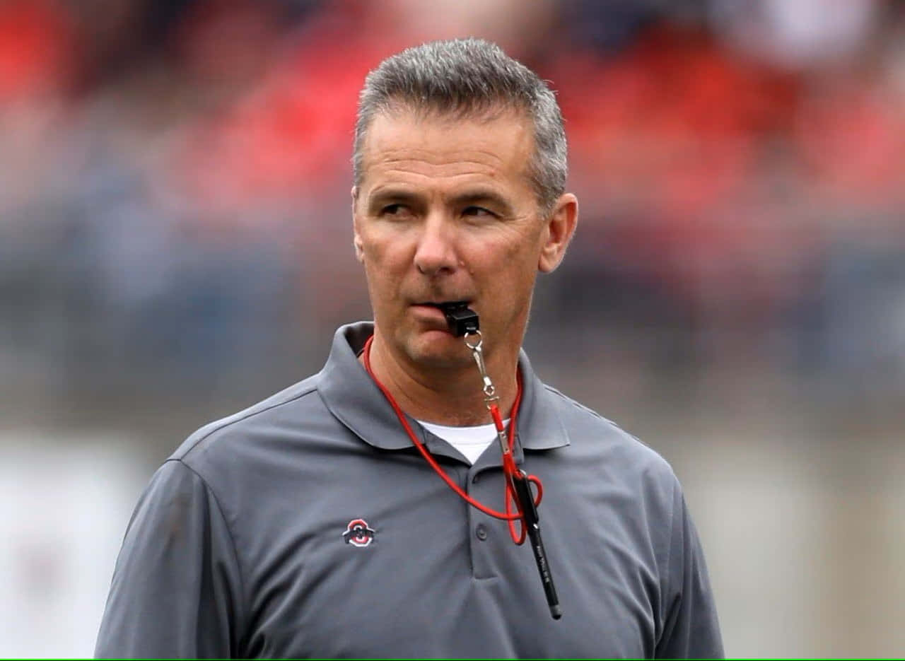 Urban Meyer 桌布 壁紙 — 一位專注的白髮足球教練，在模糊的體育場背景下將哨子放在唇邊。