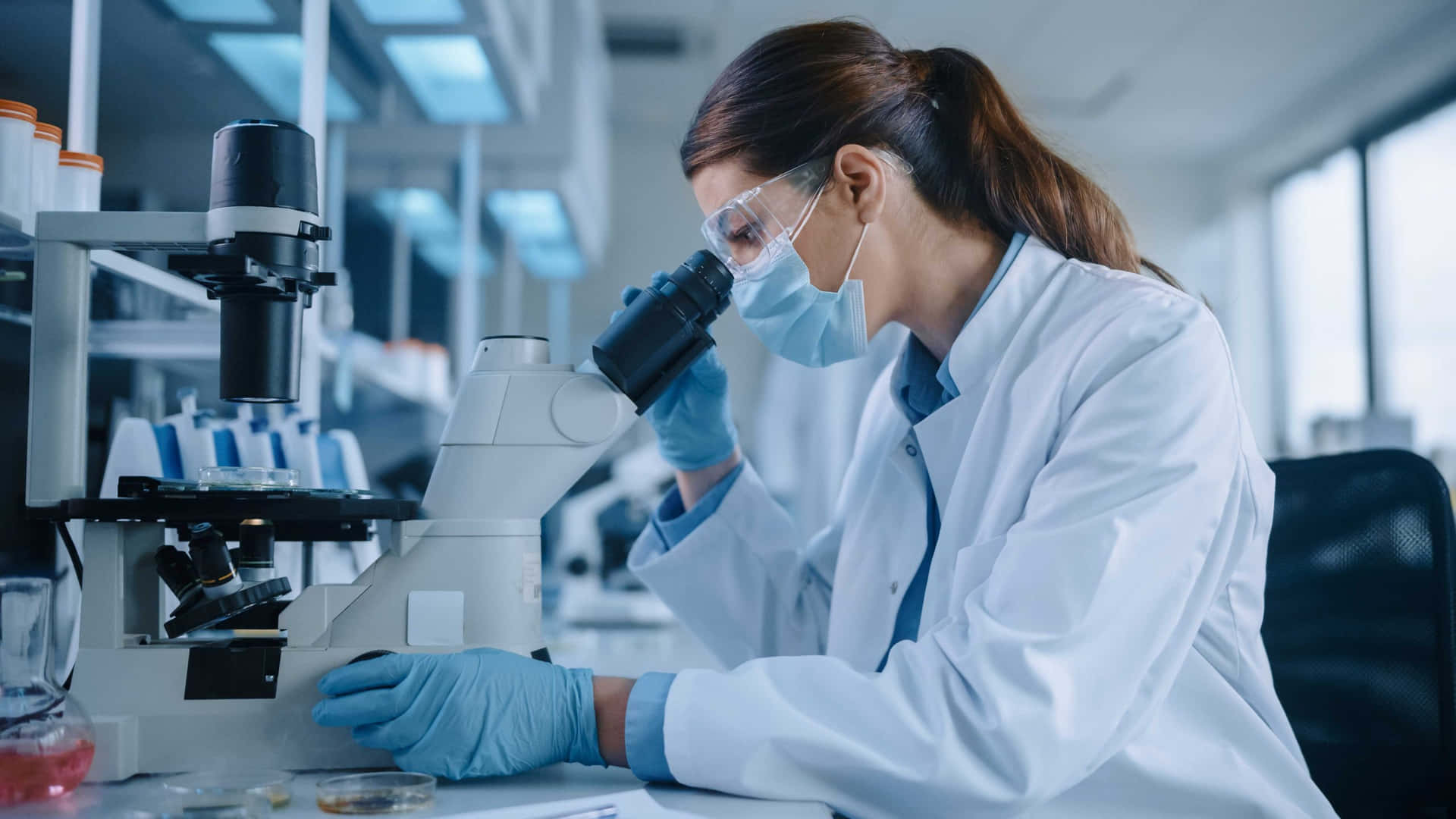 科學 桌布 壁紙 — 一位專注的科學家穿著實驗室外套和手套，在現代研究設施中透過顯微鏡觀察。