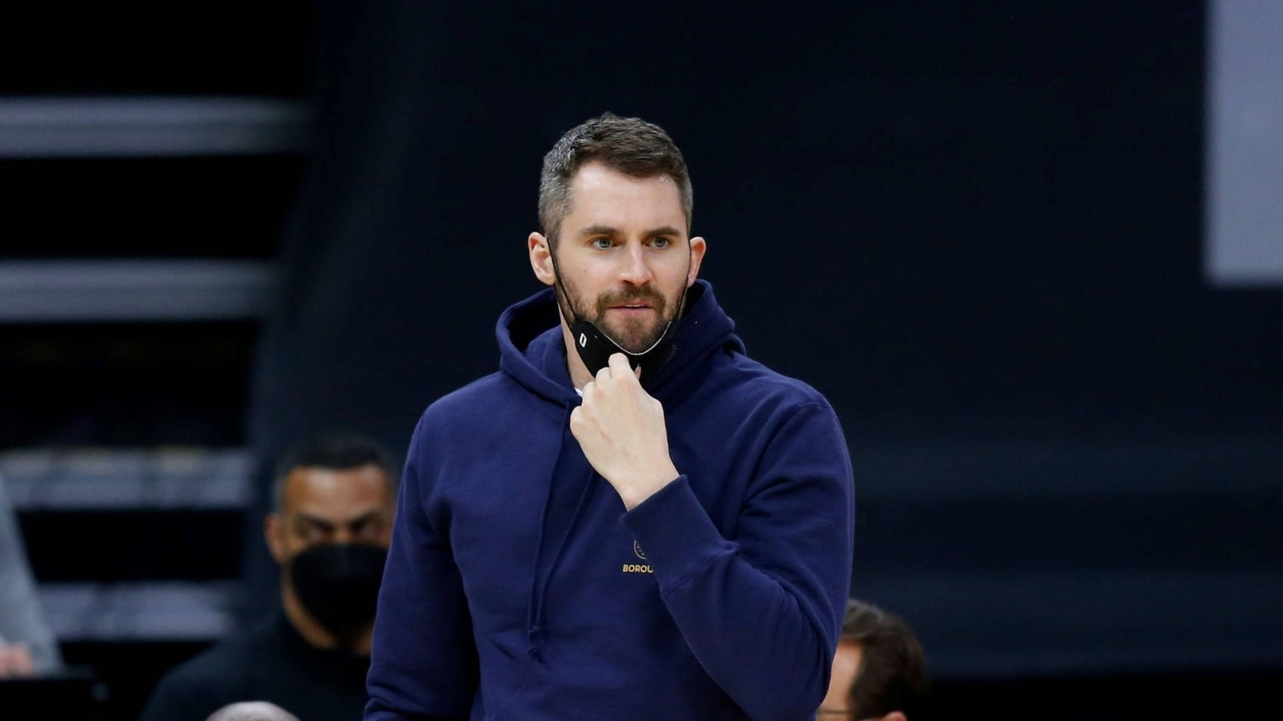 Kevin Love 桌布 壁紙 — 一位專注的籃球教練穿著海軍藍連帽衫，在明亮的場館燈光下於比賽中調整口罩。