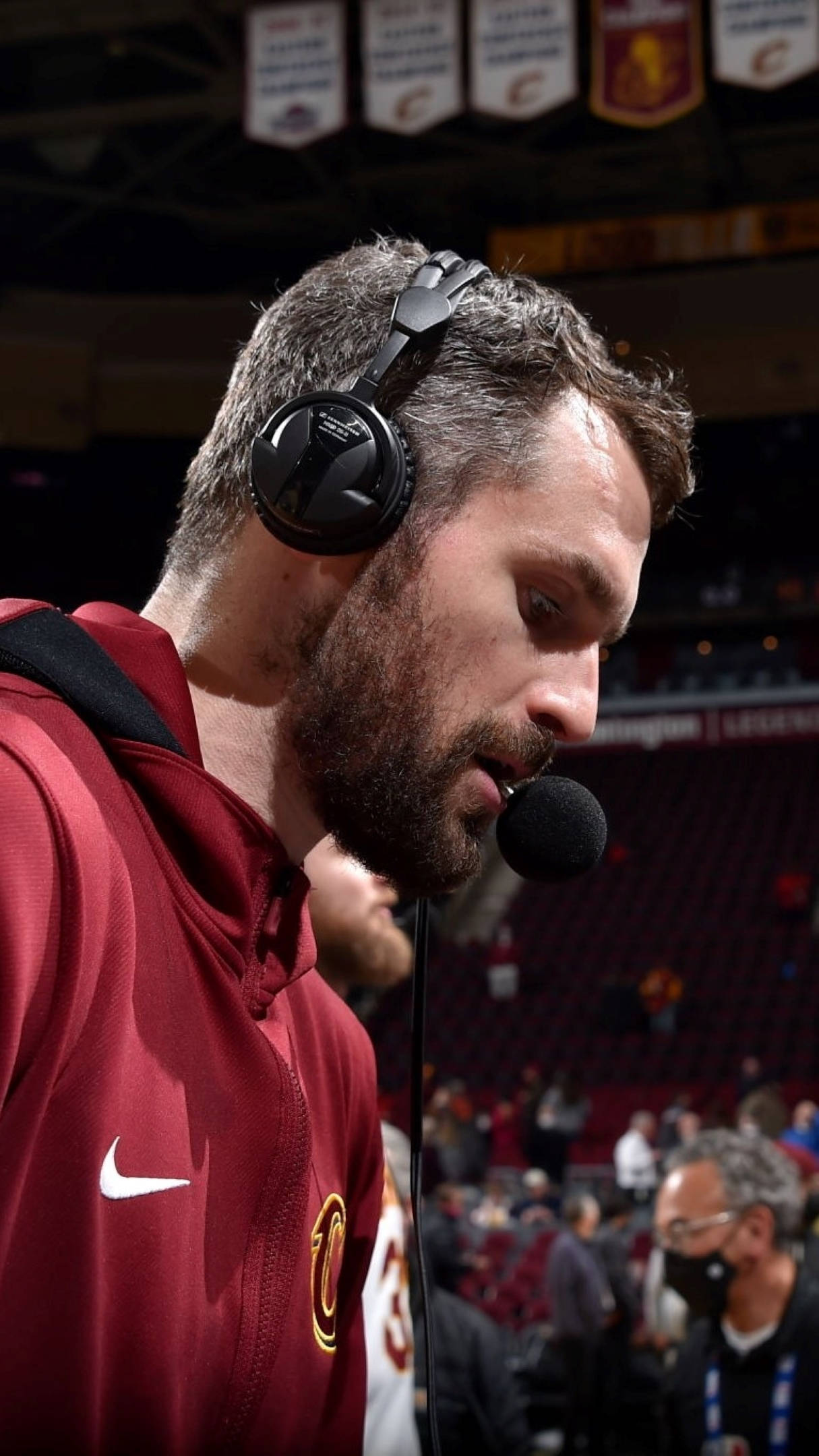 Kevin Love 桌布 壁紙 — 一位專注的籃球教練穿著酒紅色連帽衫，在擁擠的場館中戴著耳機對著麥克風說話。