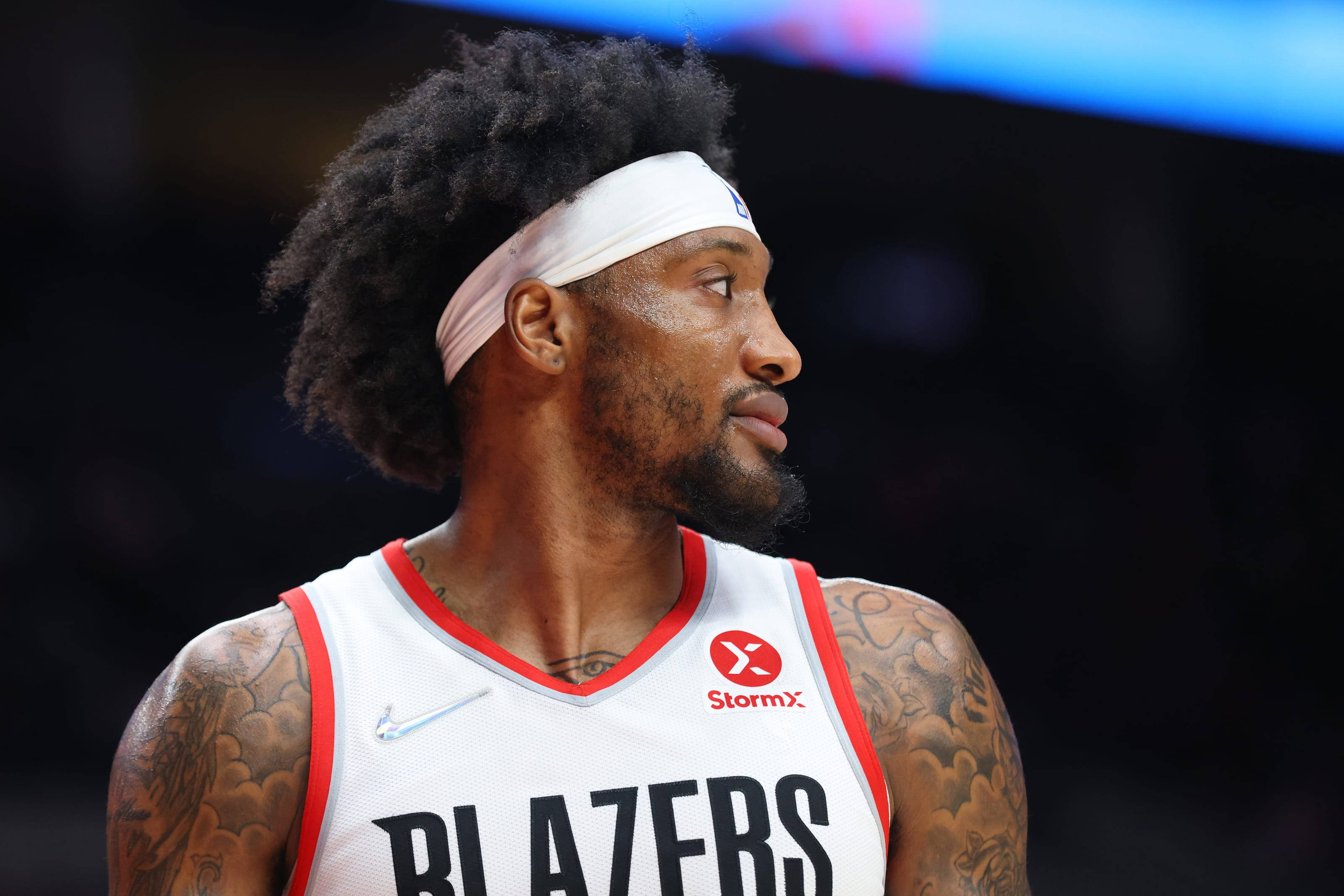 robert covington 桌布 壁紙 — 一位專注的籃球運動員，留著捲髮並有刺青，在場館燈光下穿著白色 Blazers 球衣。