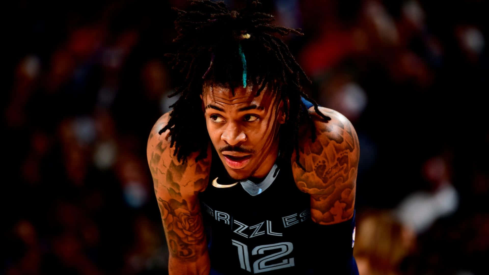 Ja Morant 動圖 桌布 壁紙 — 一位專注的籃球運動員，留著辮子頭和刺青，在黑暗的場館中於明亮燈光下向前傾身。