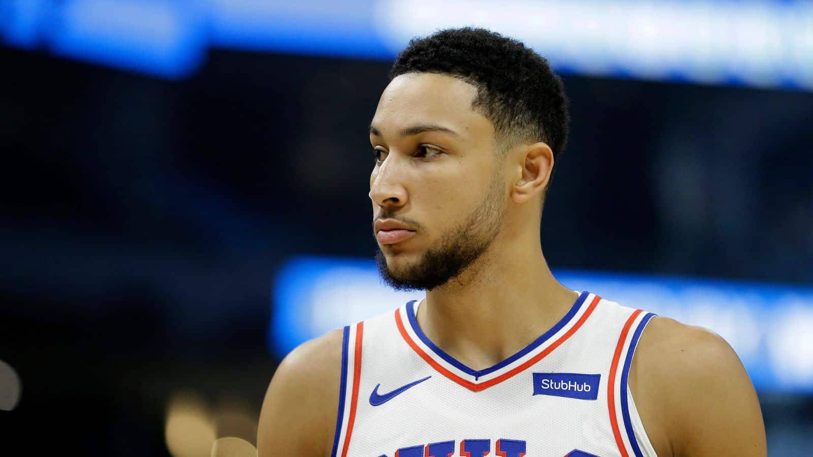 Ben Simmons 桌布 壁紙 — 一位專注的籃球運動員，身穿帶有藍色和紅色飾邊的白色球衣，站在明亮的場館燈光下的球場上。