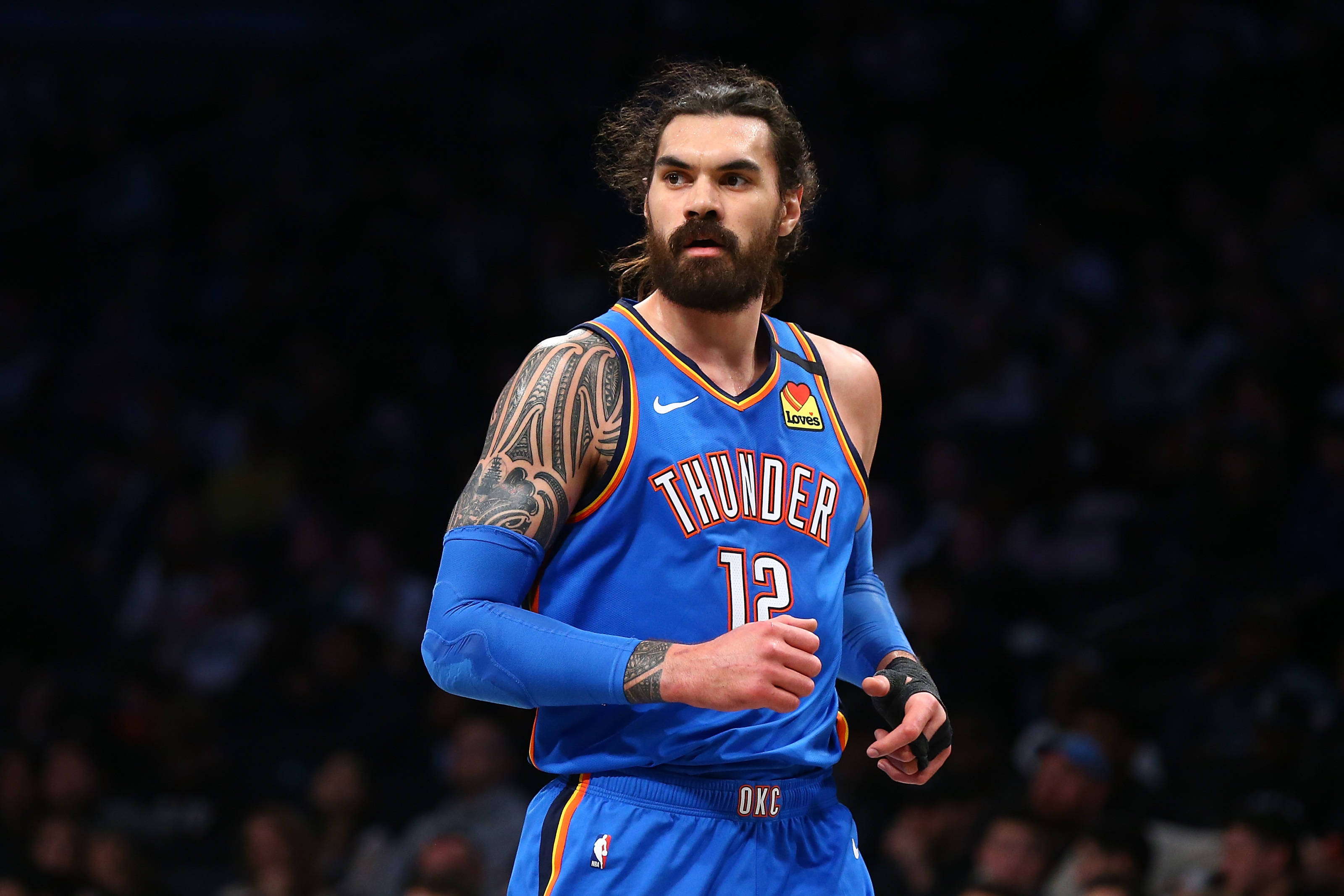 Steven Adams 桌布 壁紙 — 一位專注的籃球運動員，身穿藍色雷霆隊球衣，手臂上有刺青，站在球場上，場館燈光照射下。