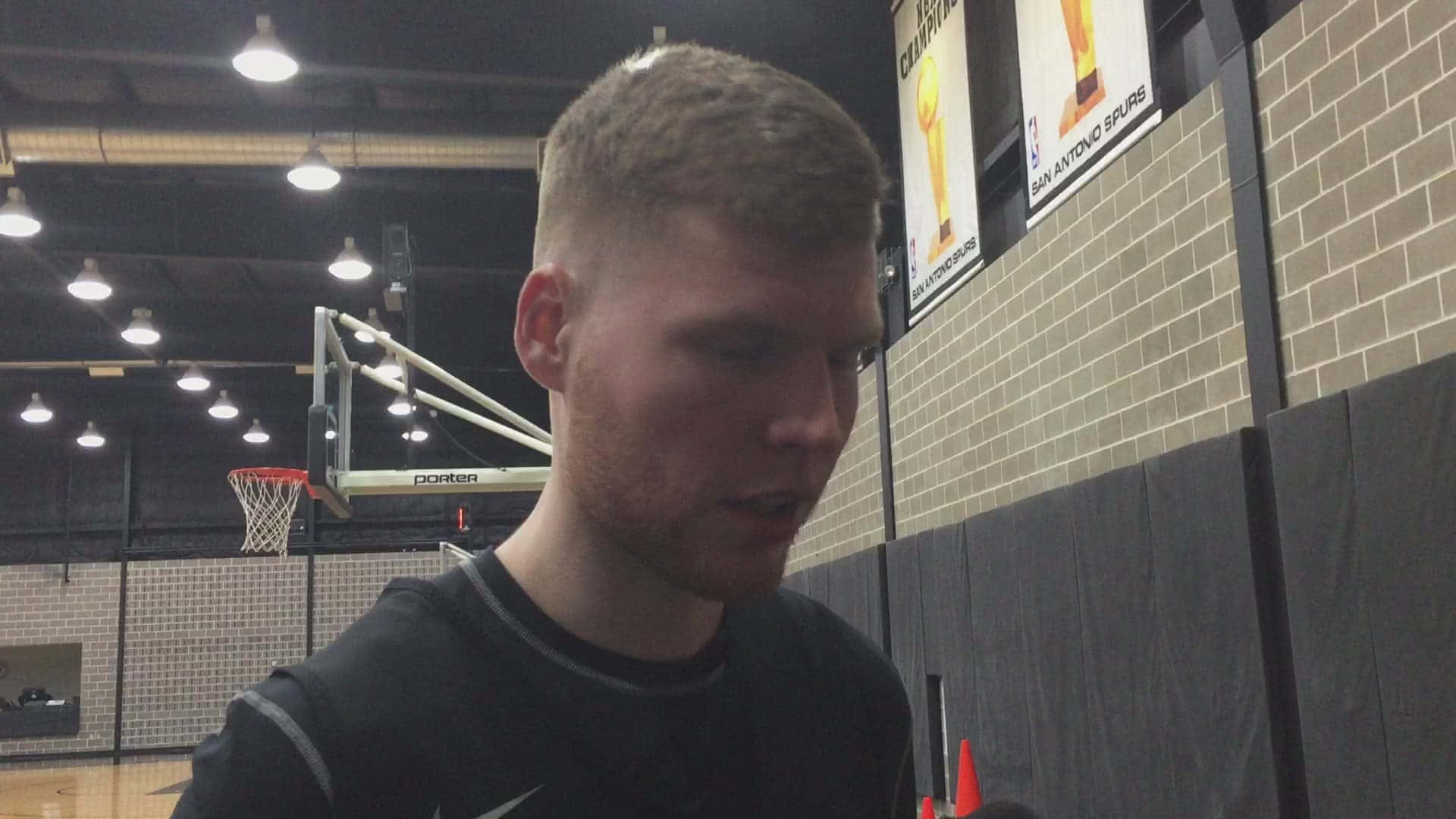 NBA 桌布 壁紙 — 一位專注的籃球運動員在健身房穿著黑色襯衫，背景有籃框和馬刺隊旗幟。