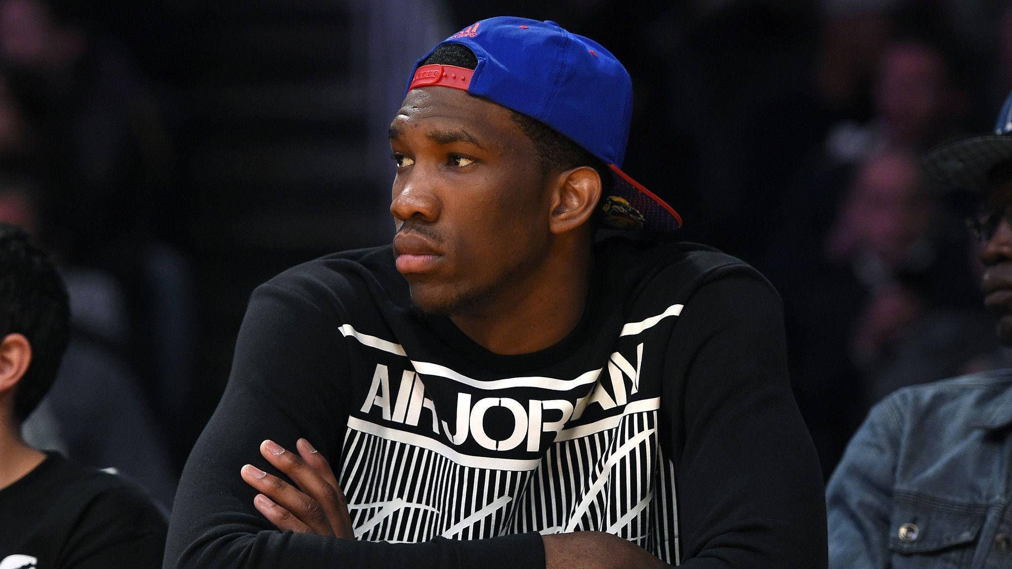 Embiid 桌布 壁紙 — 一位專注的籃球運動員戴著藍色帽子，身穿黑色Air Jordan運動衫，在比賽中交叉雙臂坐著。
