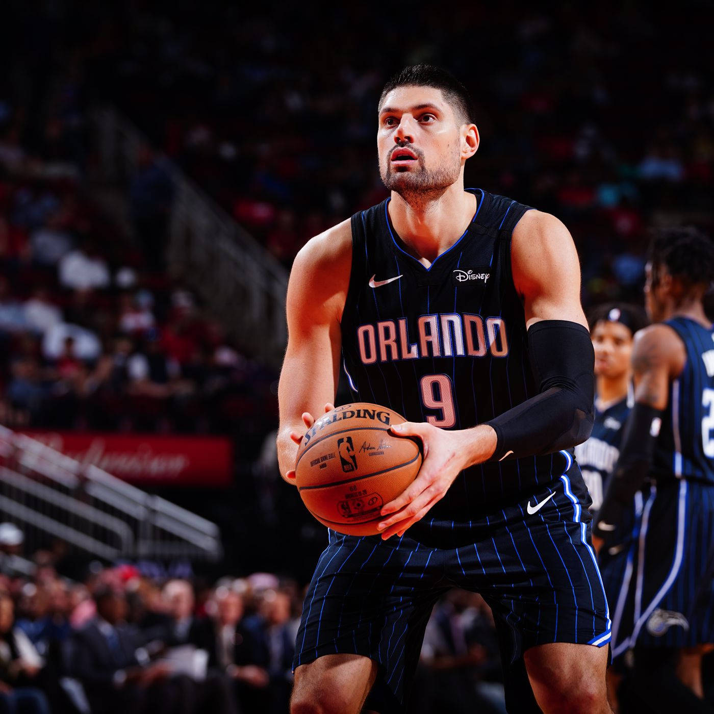 NBA 桌布 壁紙 — 一位專注的籃球運動員穿著奧蘭多球衣，在球場燈光下比賽中持球，背景有模糊的觀眾。