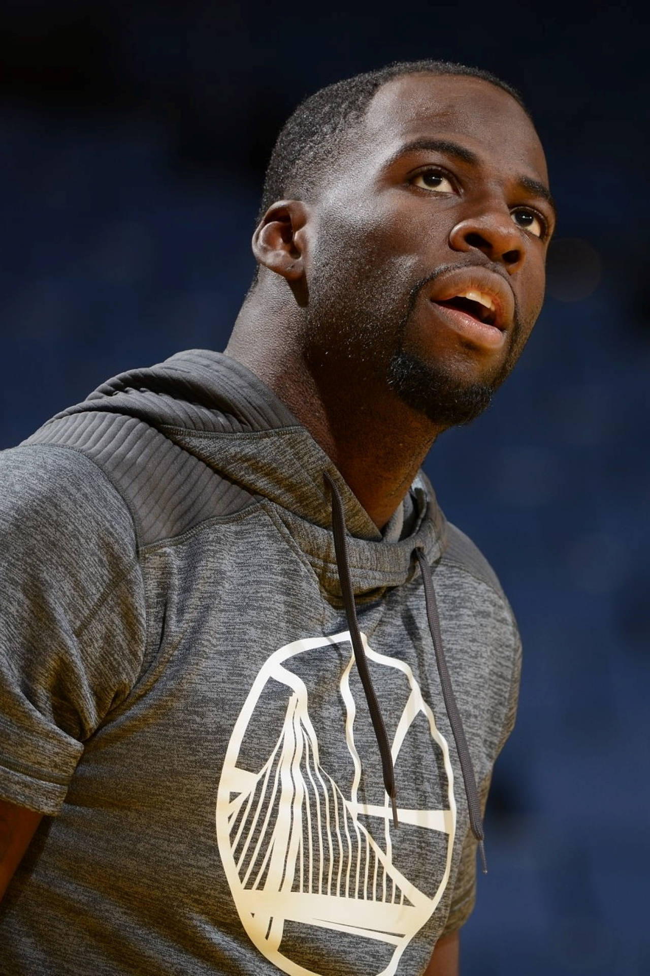 Draymond Green 桌布 壁紙 — 一位專注的籃球運動員穿著灰色連帽衫，上面有金色球隊標誌，在場館燈光下比賽時向上看。