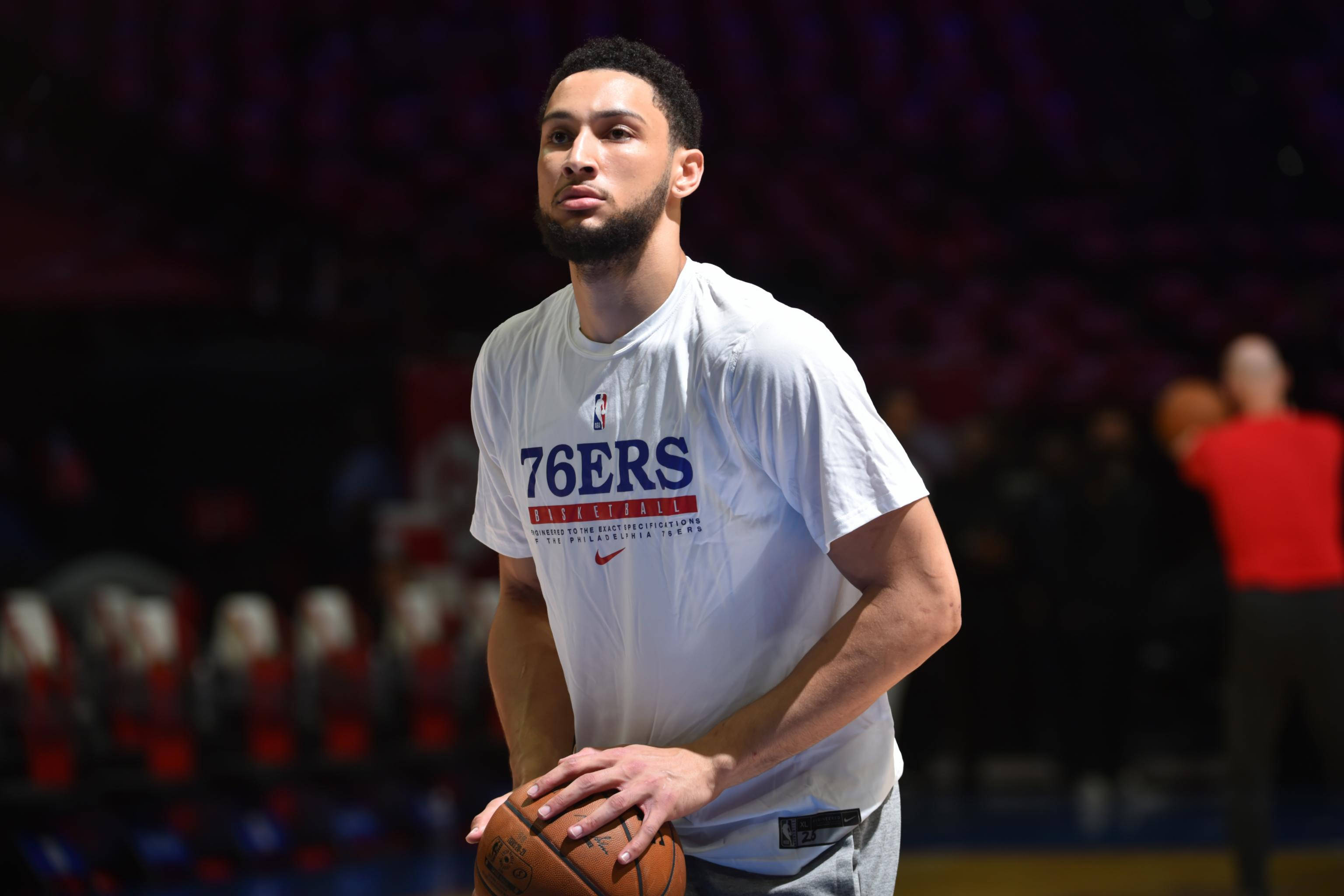 76ers 桌布 壁紙 — 一位專注的籃球運動員穿著白色76ers練習衫，在場館燈光下拿著球，身後是模糊的紅色座位。