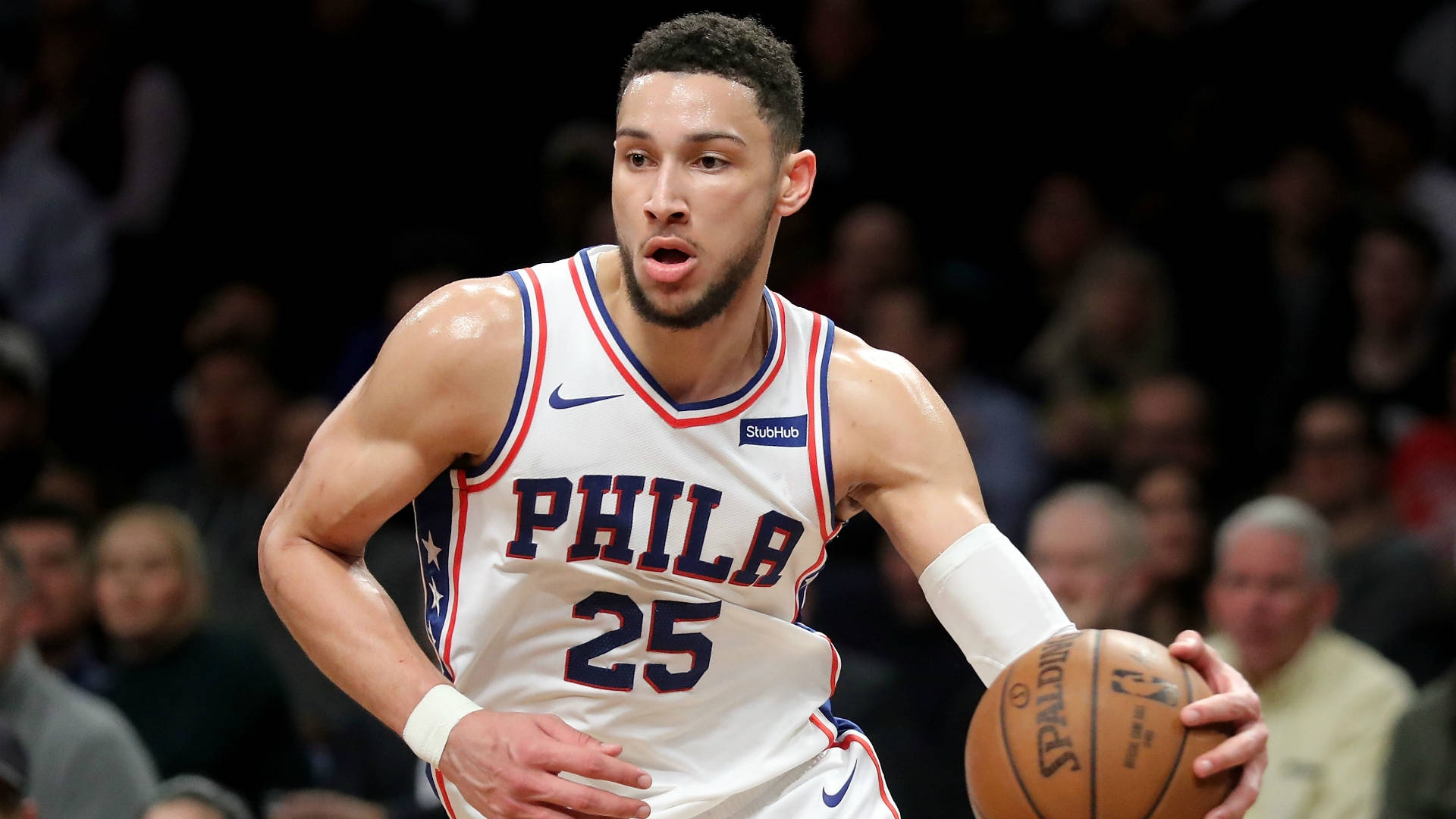 Ben Simmons 桌布 壁紙 — 一位專注的籃球運動員穿著白色Phila球衣，在明亮的場館燈光下於激烈比賽中運球。