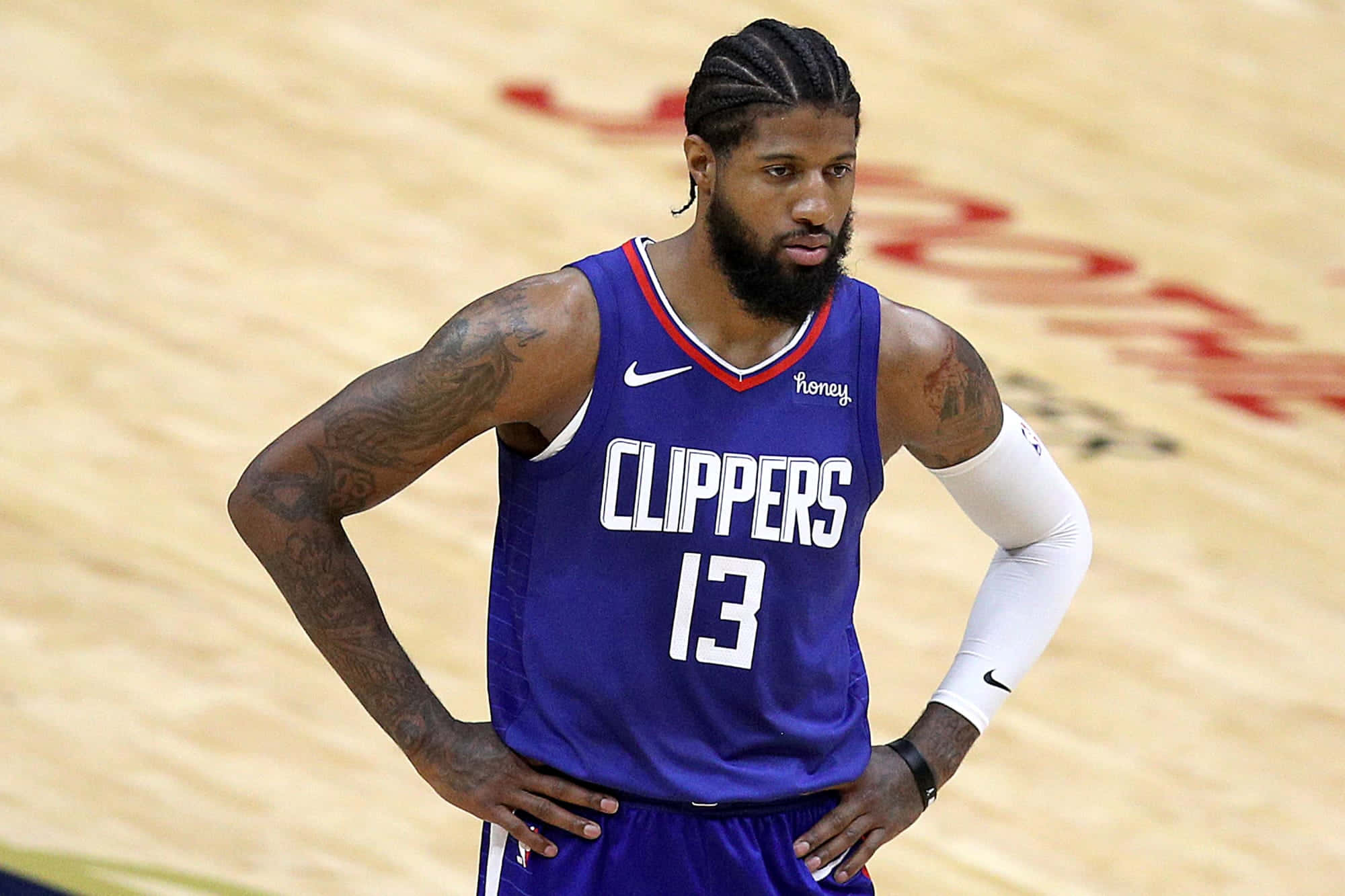 Paul George Clippers 桌布 壁紙 — 一位專注的籃球運動員穿著紫色快艇隊球衣，雙手叉腰站在木地板上。