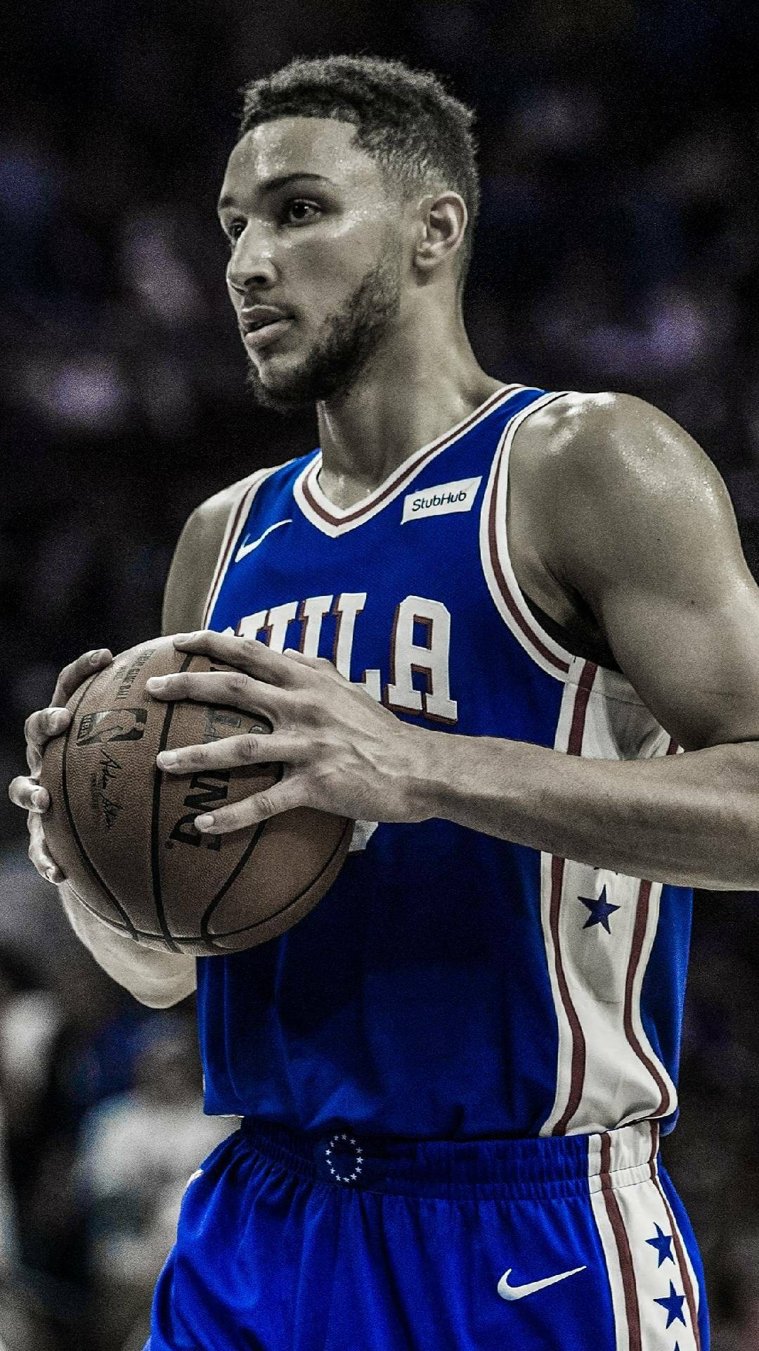 Ben Simmons 桌布 壁紙 — 一位專注的籃球運動員穿著藍色費城球衣，在模糊的人群背景的體育館燈光下拿著球。