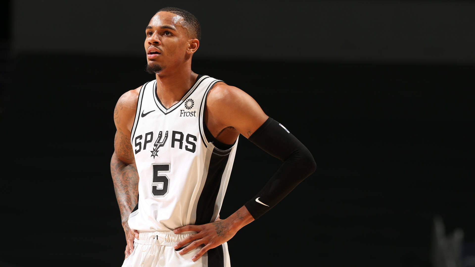 Dejounte Murray 桌布 壁紙 — 一位專注的籃球運動員身穿Spurs球衣，雙手叉腰站在深色背景前。