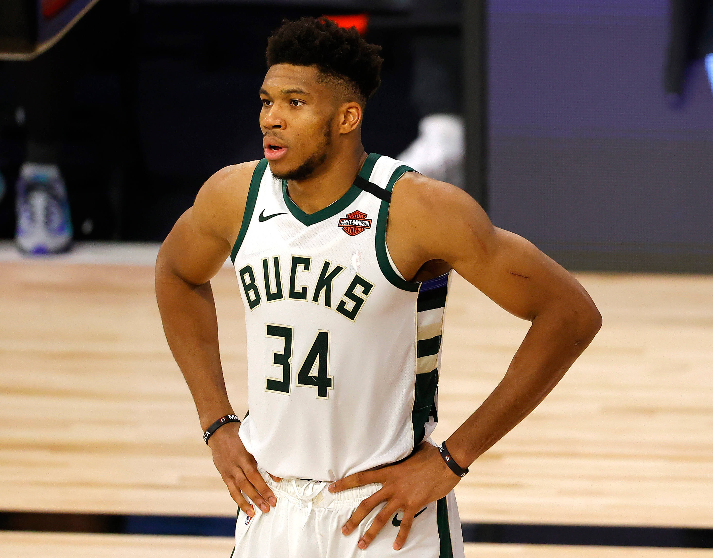 Giannis 桌布 壁紙 — 一位專注的籃球運動員身穿公鹿隊球衣，在明亮的場館燈光下，雙手叉腰站在拋光球場上。
