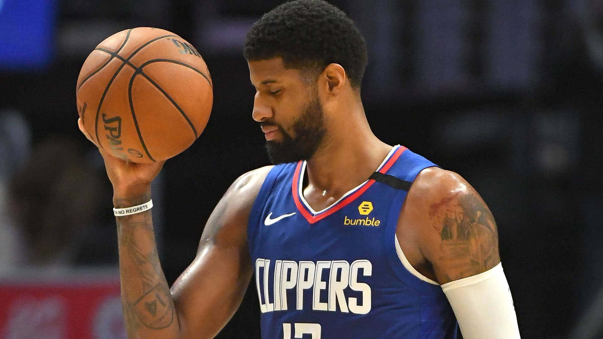 Paul George Clippers 桌布 壁紙 — 一位專注的籃球運動員身穿快艇隊球衣，在場館燈光下持球，手臂上的刺青清晰可見。