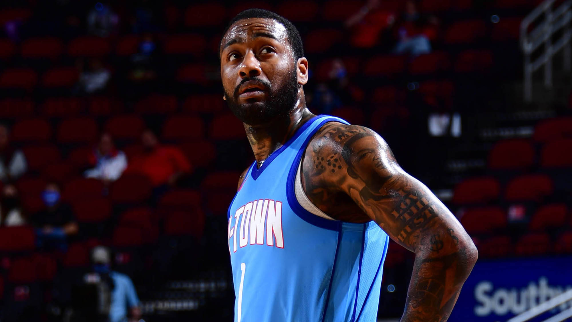 John Wall 桌布 壁紙 — 一位專注的籃球運動員身穿淺藍色球衣，手臂上有刺青，站在背景有紅色座位的球場上。