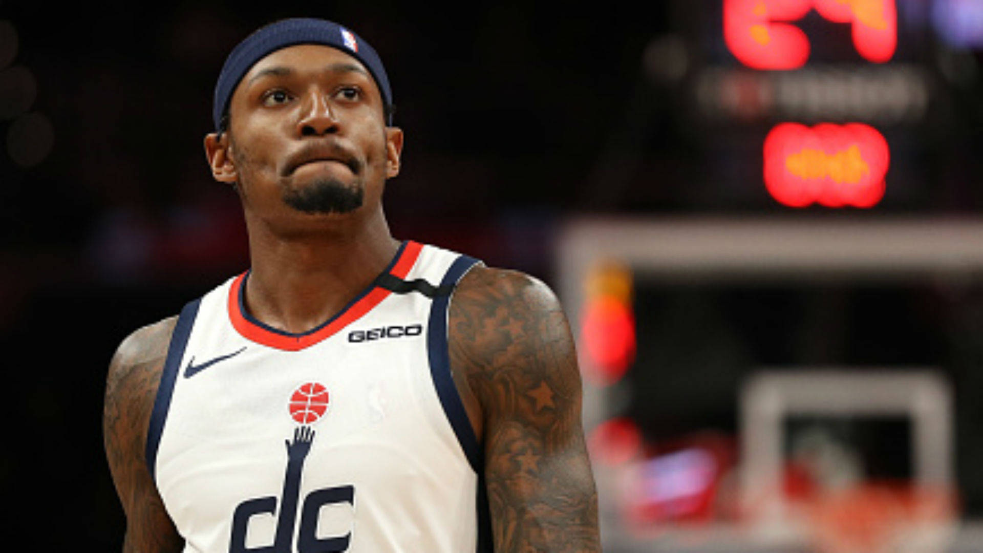 Bradley Beal 桌布 壁紙 — 一位專注的籃球運動員身穿白色DC球衣，帶有刺青，在比賽期間於場館燈光下向上凝視。