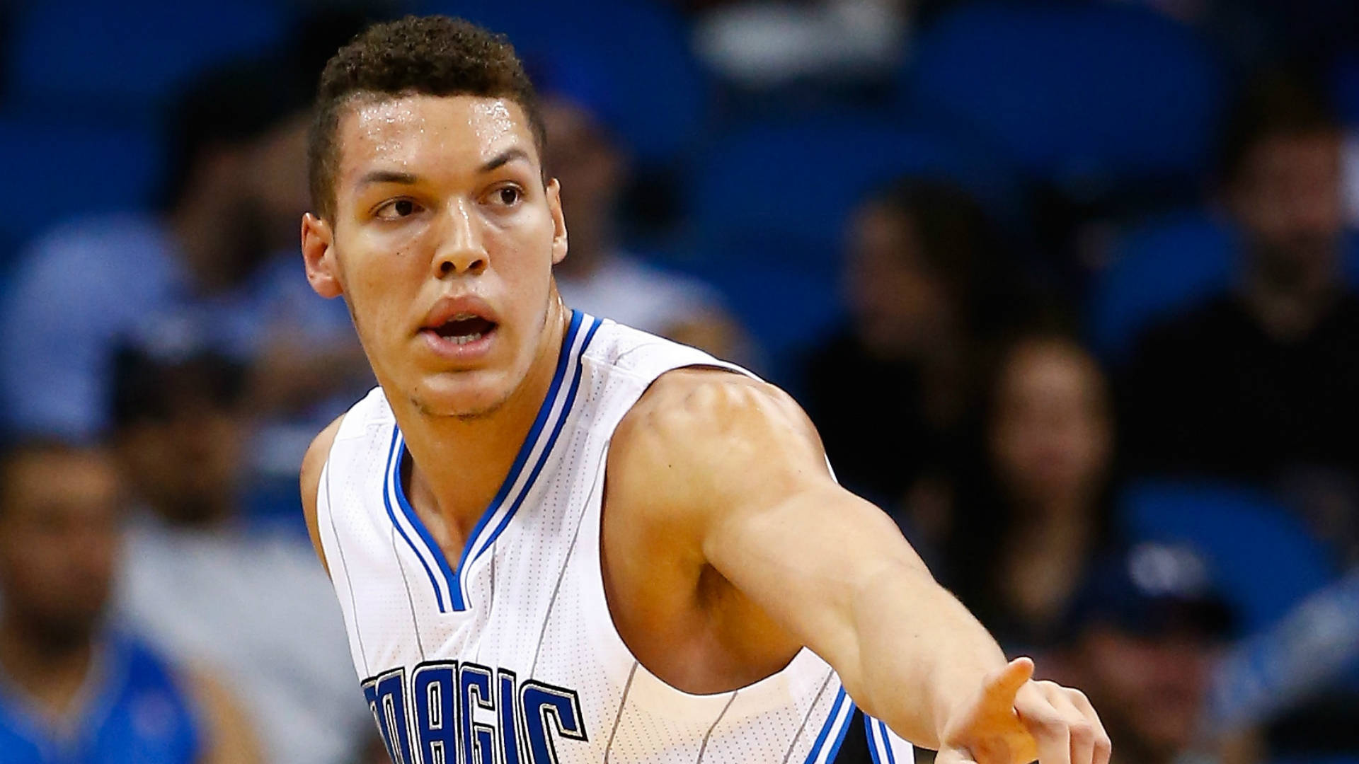 aaron gordon 桌布 壁紙 — 一位專注的籃球運動員身穿白色奧蘭多魔術隊球衣，在模糊的藍色體育場座位背景下激烈運球。
