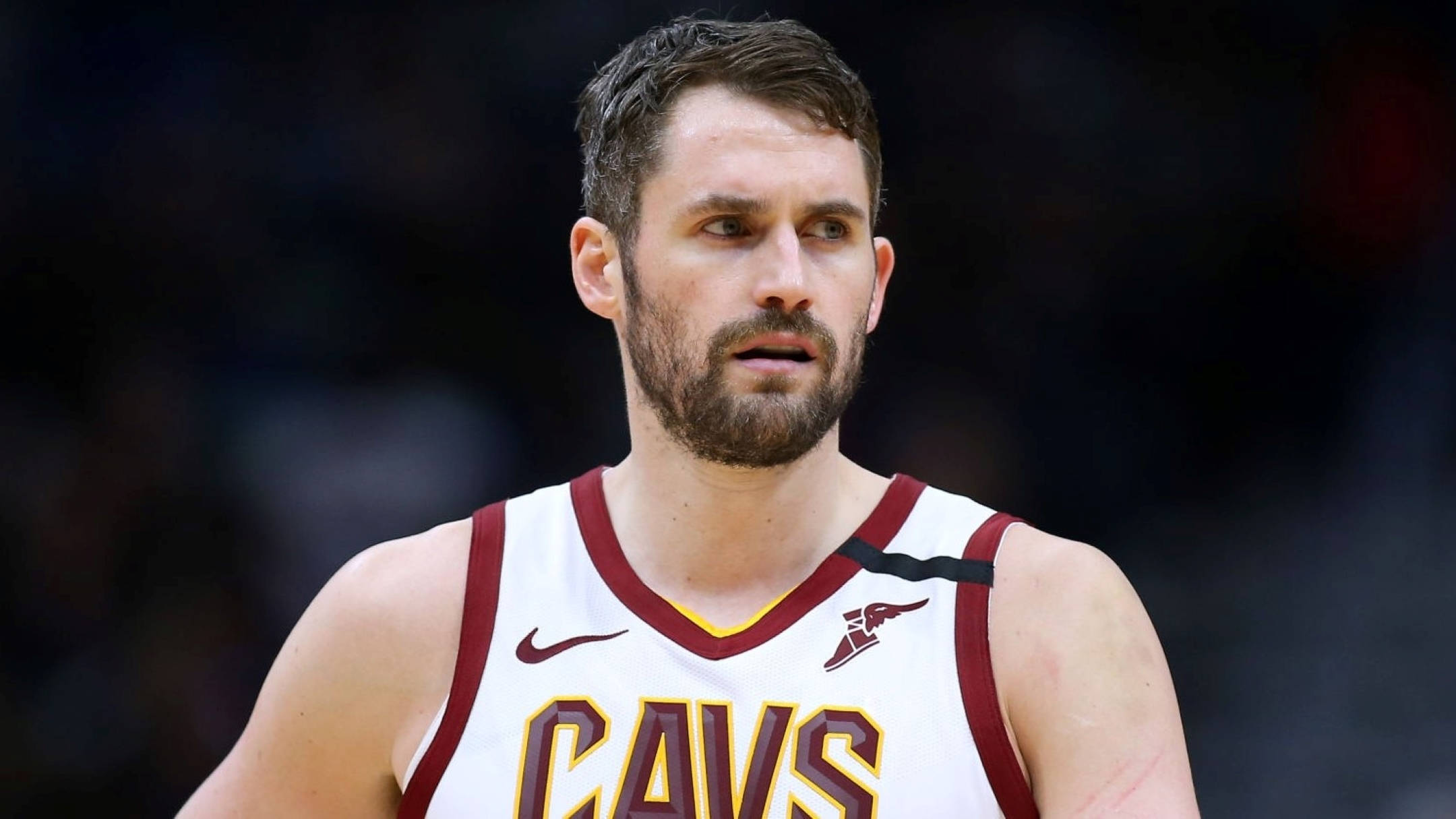Kevin Love 桌布 壁紙 — 一位專注的籃球運動員身穿白色栗色Cavs球衣，在明亮的場館燈光下於比賽中凝視著。