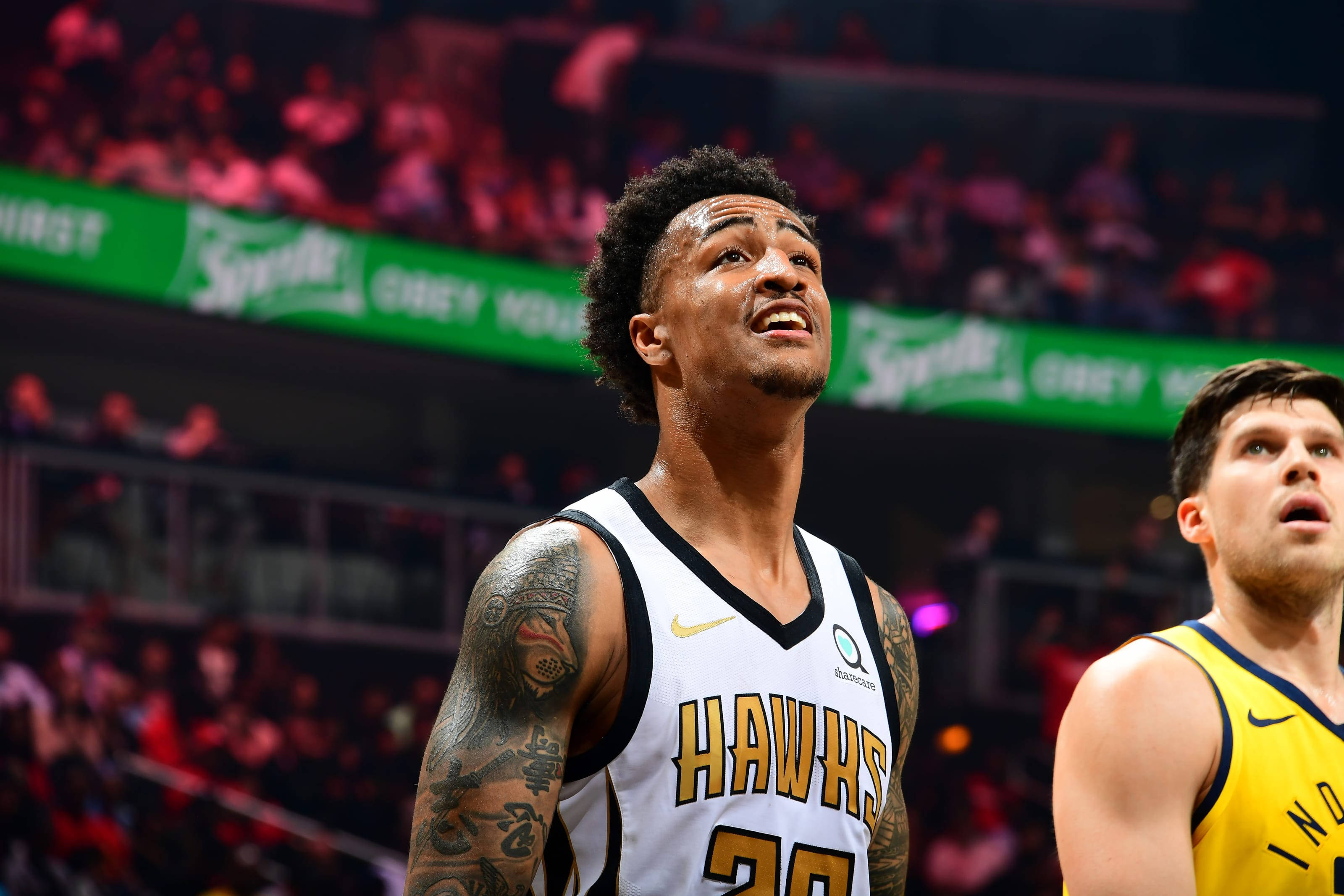 Hawks 桌布 壁紙 — 一位專注的籃球運動員身穿白色老鷹隊球衣，帶有刺青，在場館燈光下激烈的比賽中向上凝視。