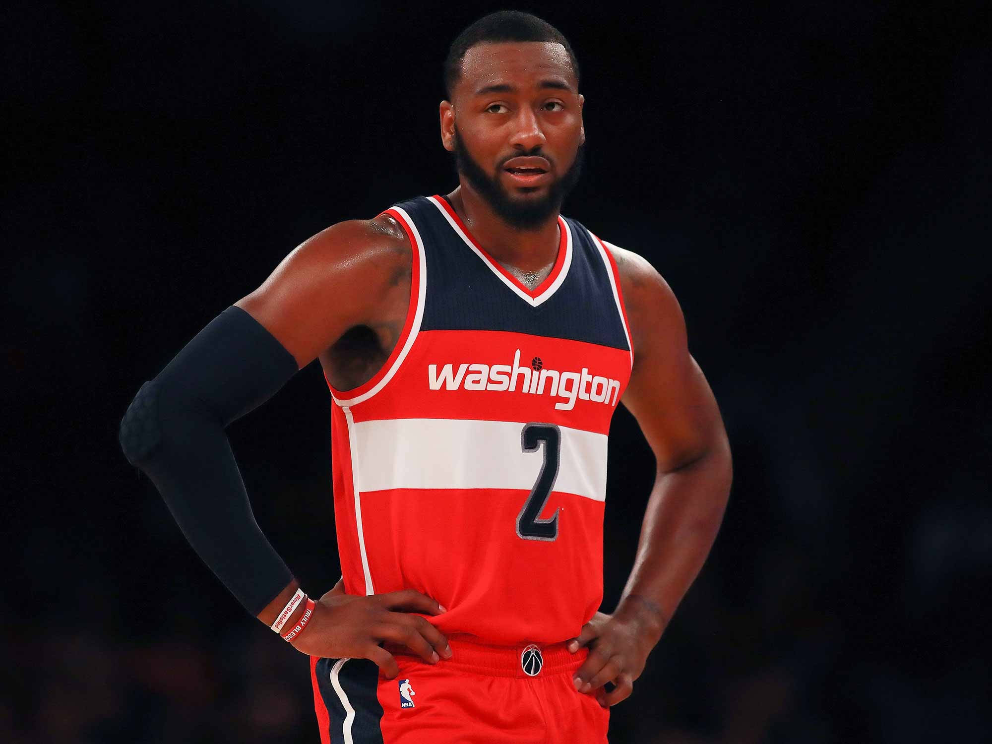 John Wall 桌布 壁紙 — 一位專注的籃球運動員身穿紅白藍華盛頓球衣，背號二號，雙手叉腰站在黑暗的球場背景下。