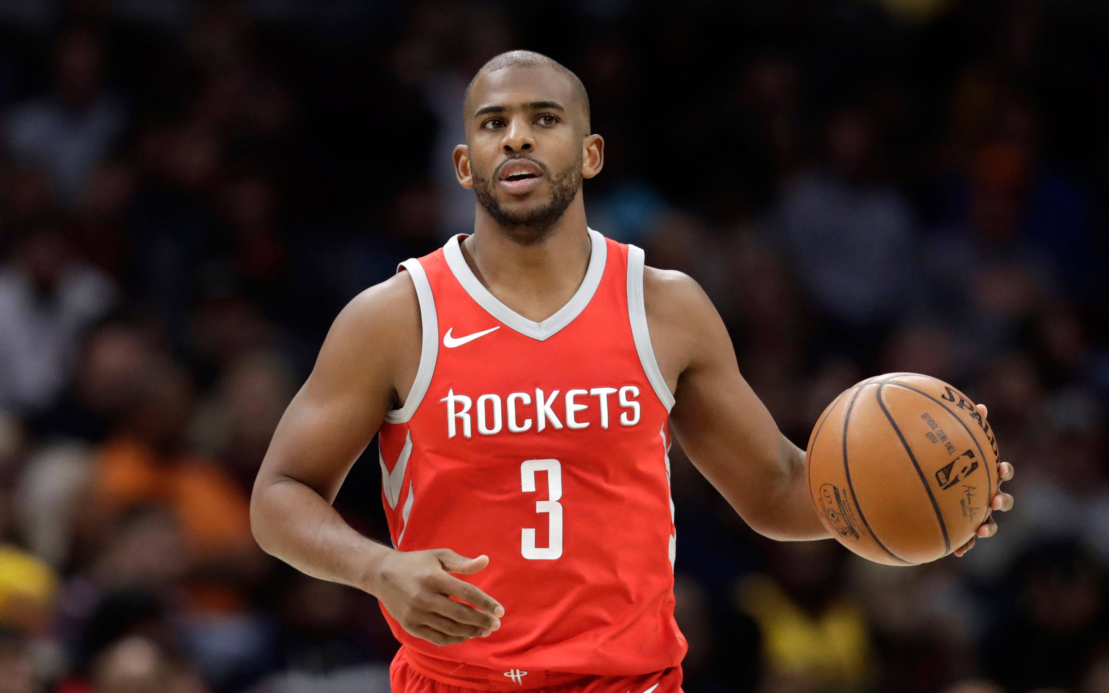 Chris Paul 桌布 壁紙 — 一位專注的籃球運動員身穿紅色火箭隊球衣，在明亮的場館燈光下於激烈比賽中運球。